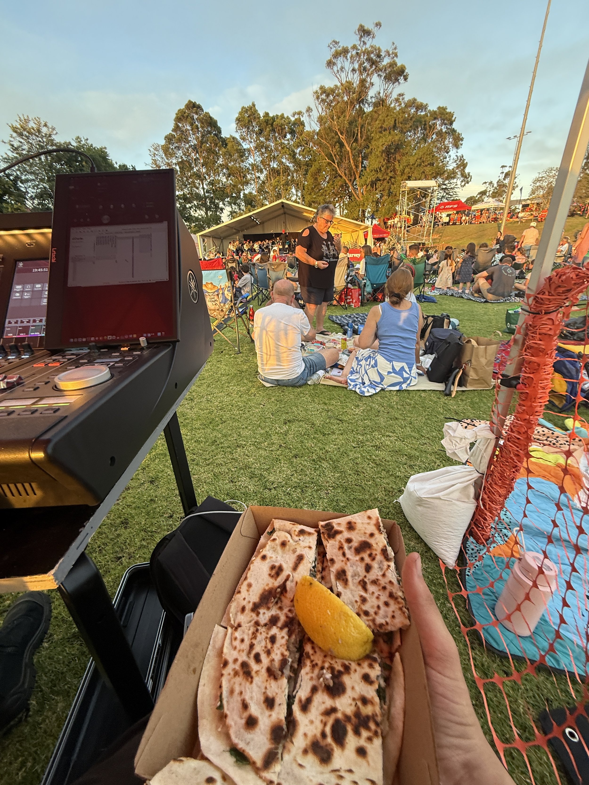 A person holds a box of grilled quesadilla sandwiches with lemon wedge in the foreground at an outdoor concert or festival. In the background, people are sitting on the grass, some on blankets, enjoying the event near a stage with trees and sky overh