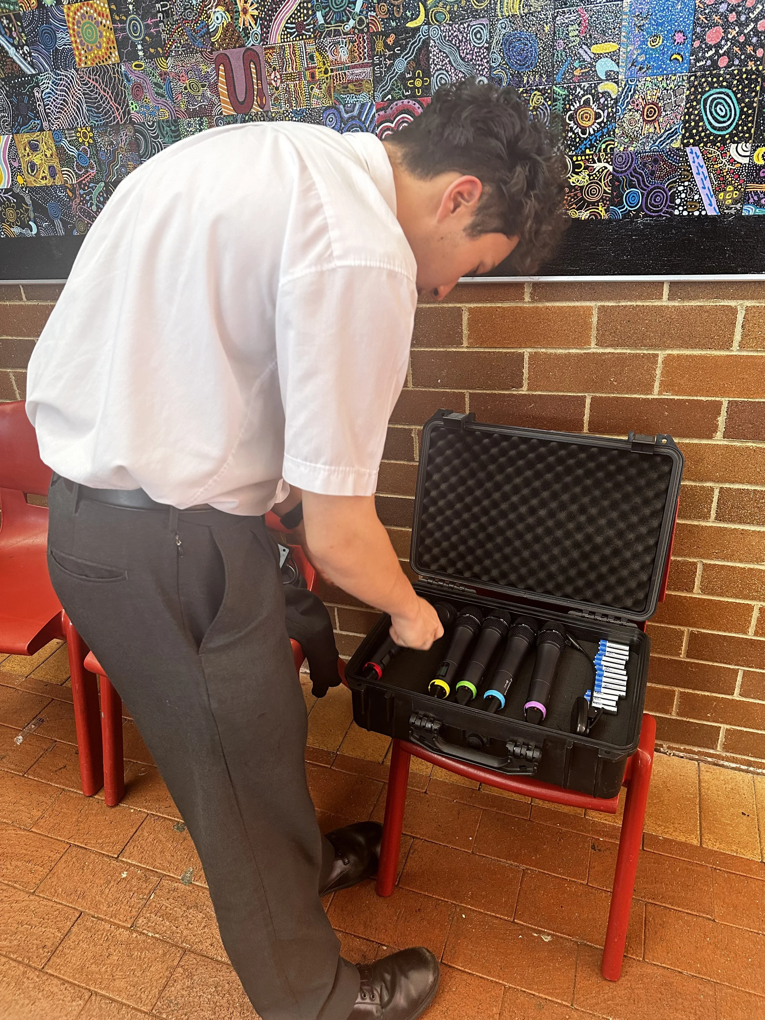 A man with dark curly hair wearing a white shirt and dark trousers is opening a black carrying case with microphones and batteries inside, placed on a red chair. The background features a brick wall and a colorful abstract art piece.