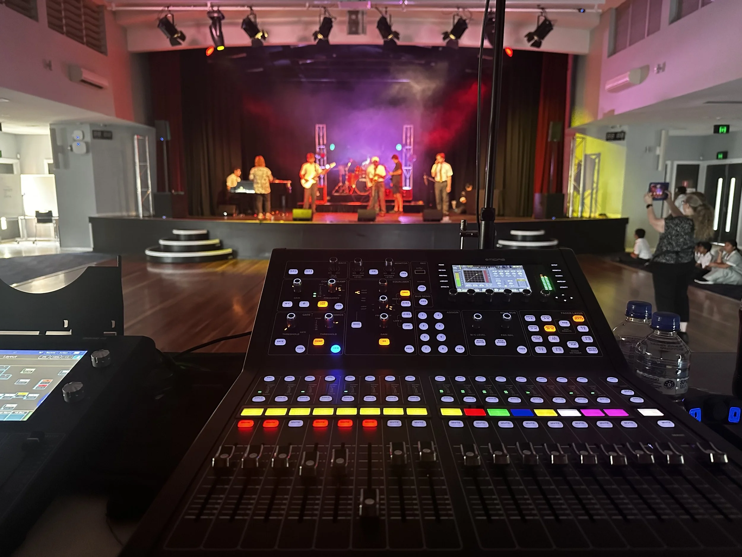 View from the sound mixing console showing a stage with a live band performing, colorful stage lights, and audience members, including a woman taking a photo, watching the show.