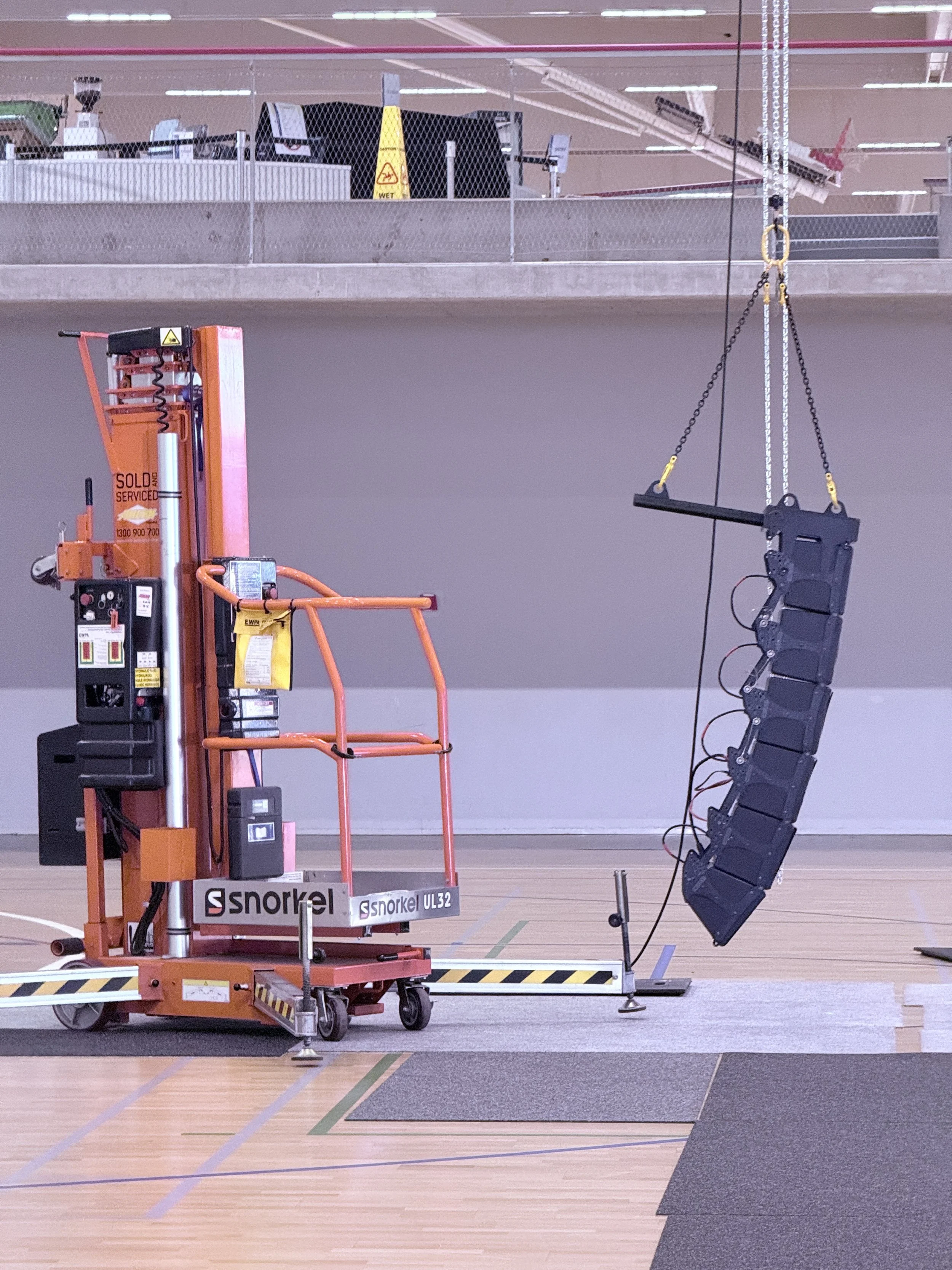 A mobile lift with a speaker system hanging from the ceiling of an indoor sports gymnasium.