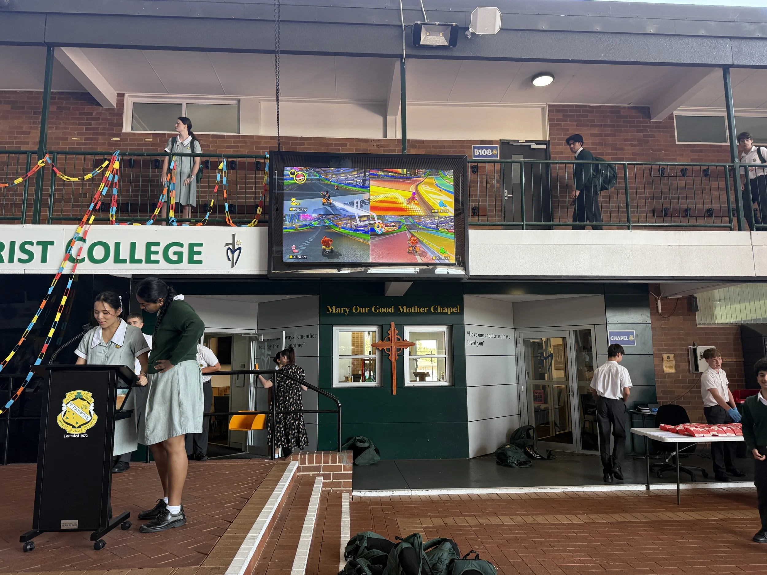 Students at a school event, some playing a video game on a large screen, others standing and talking outside the Mary Our Good Mother Chapel at a Christian college.