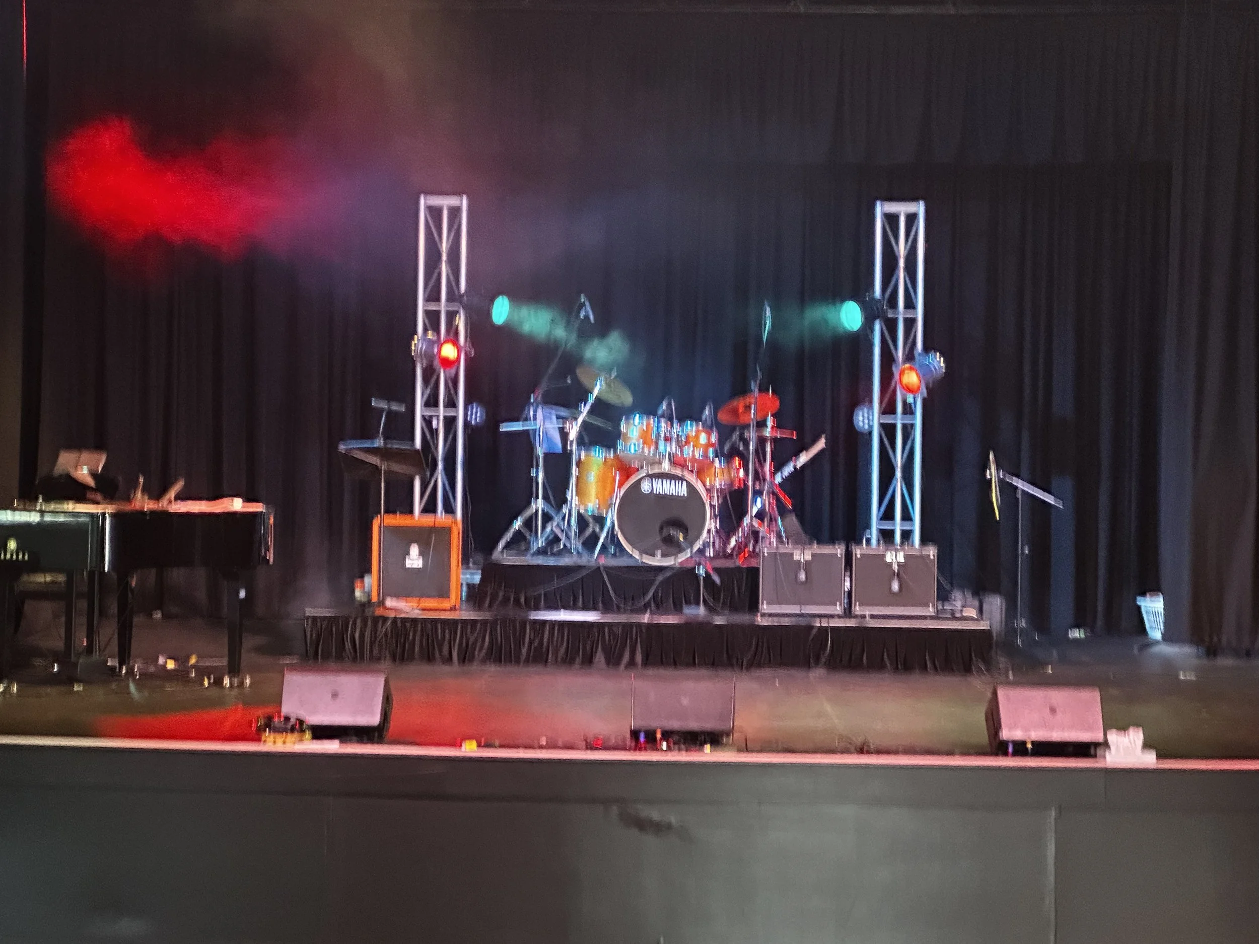 Empty stage with musical instruments including a drum set, keyboard, and amplifier, with colorful stage lights and a black curtain backdrop.