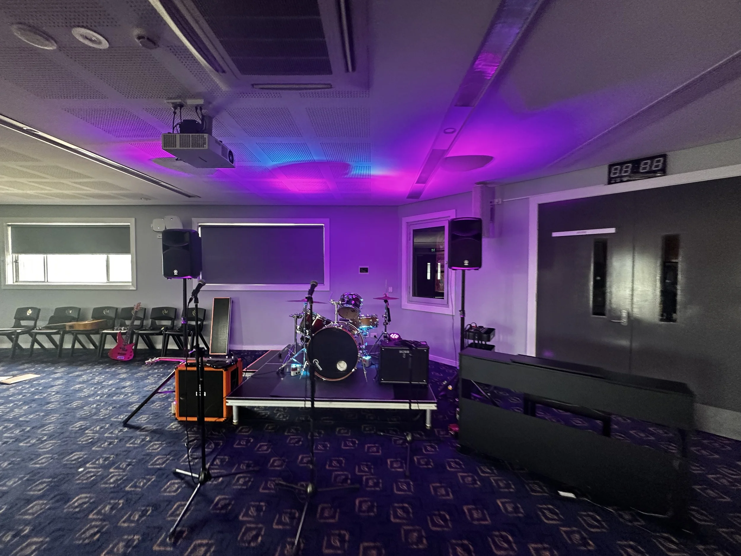Concert stage set up with drums, microphones, and amplifiers in a dimly lit room with purple and pink lighting.