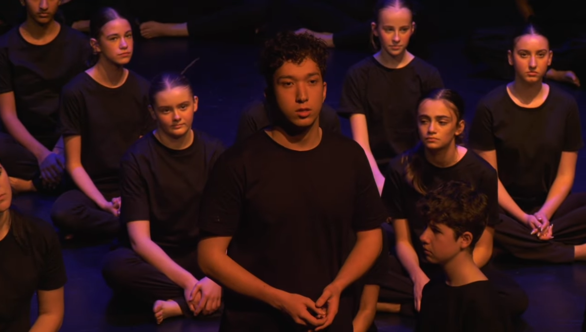 Group of children sitting cross-legged on the floor, wearing black shirts, during a performance or rehearsal.