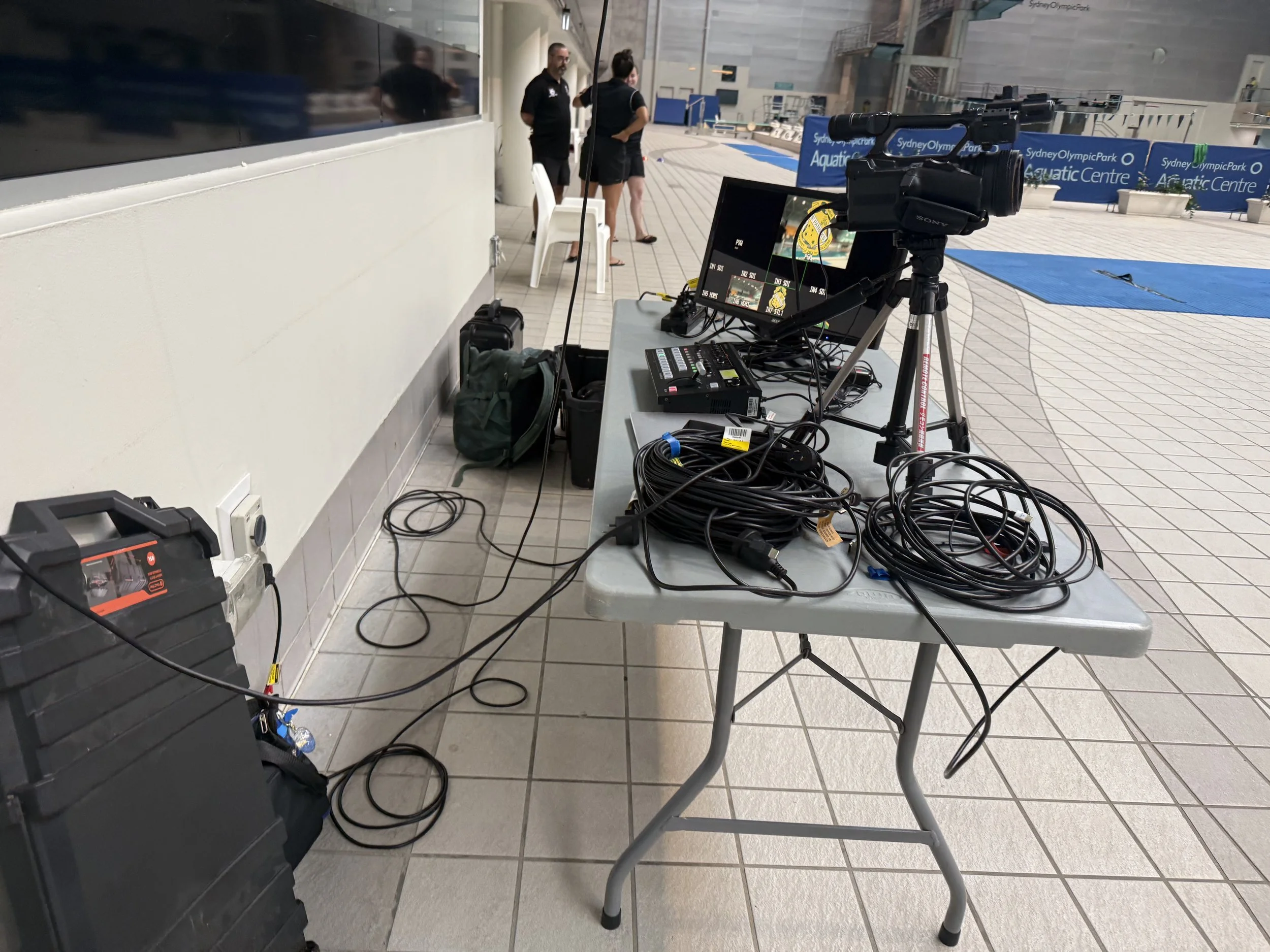 A table with electronic equipment including a camera on a tripod, numerous cables, and monitors at an indoor aquatic center with people visible in the background.