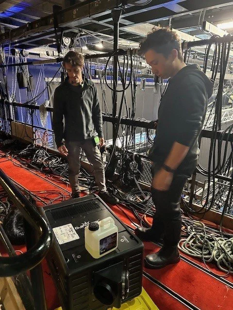 Two men in backstage area surrounded by cables and equipment, with one operating a fog machine.