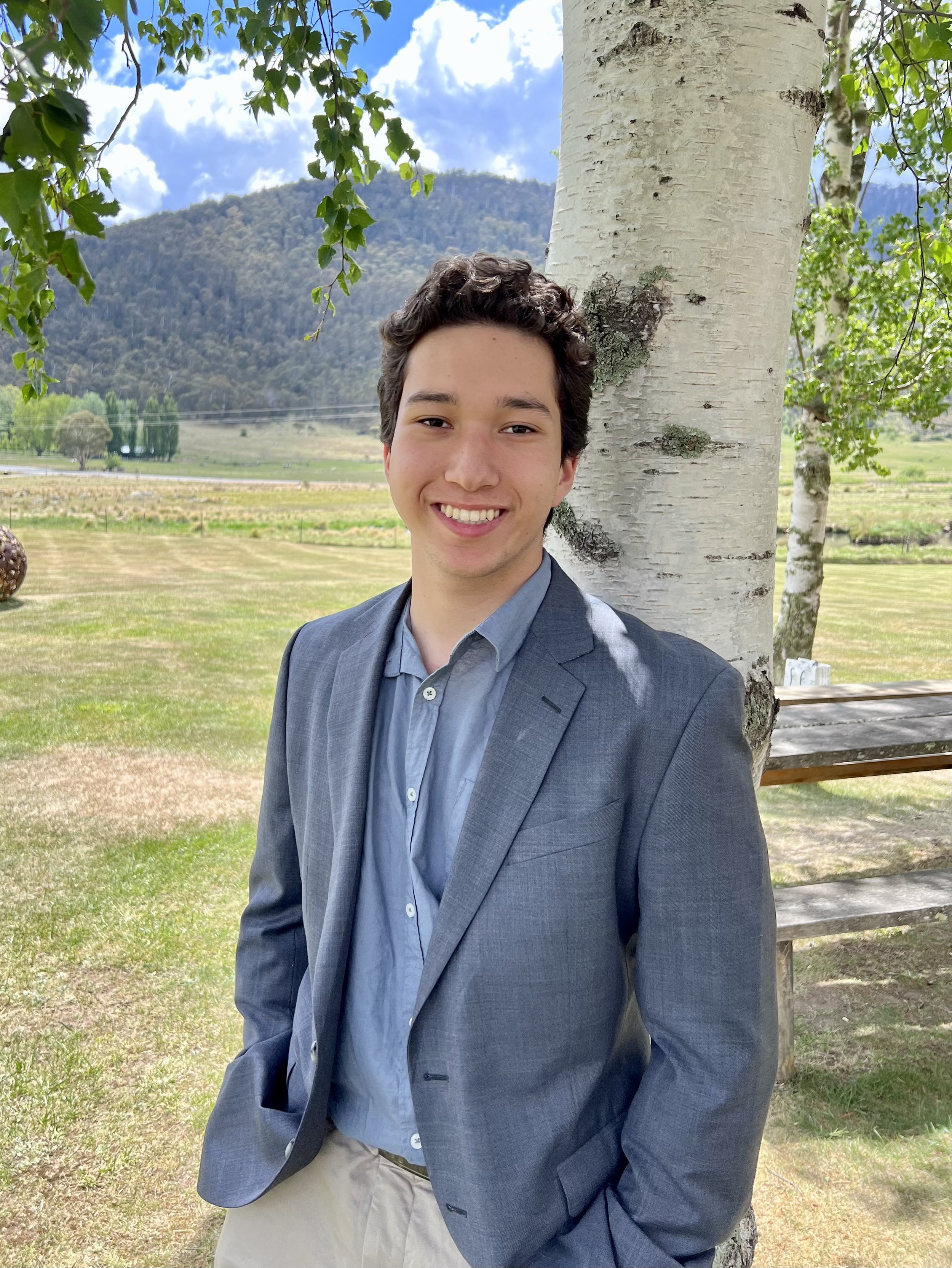 A young man in a gray suit jacket and light-blue shirt stands outdoors near a large tree, with a mountain and cloudy sky in the background.