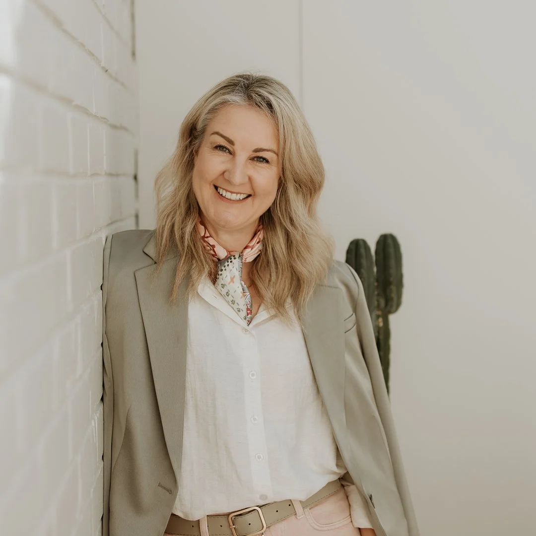 Smiling woman with shoulder-length black hair wearing a blue top, outdoors with green foliage in the background.