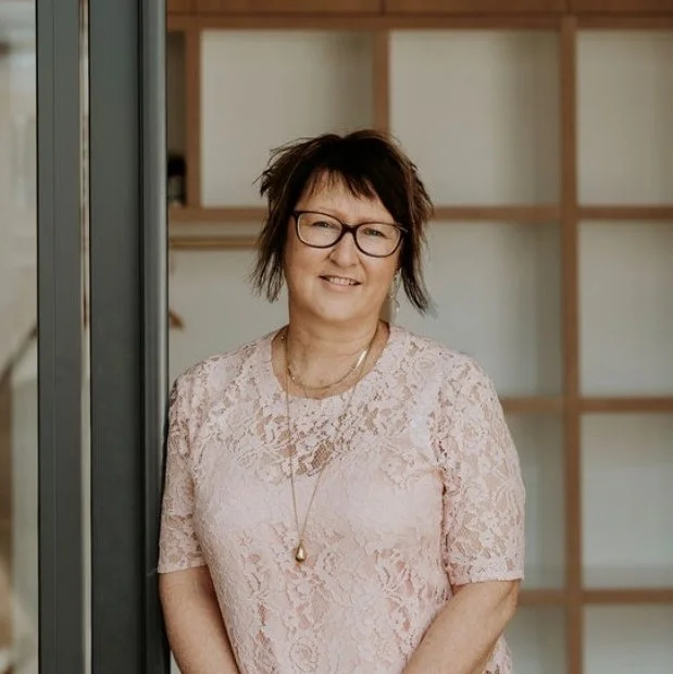 A man with dark, curly hair, glasses, and a beard, wearing a black sweater, sitting indoors with a neutral background and a plant in the blurred background.
