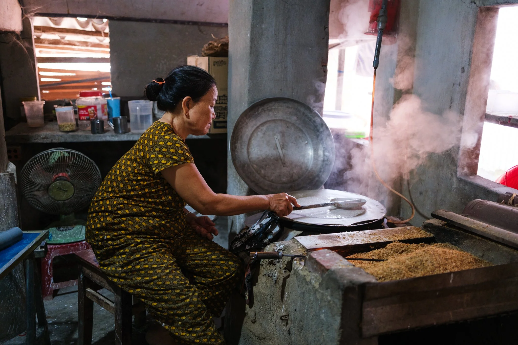 A woman in traditional patterned clothing is cooking on a stove in a rustic kitchen with steam rising, various containers on shelves, and a fan in the background.