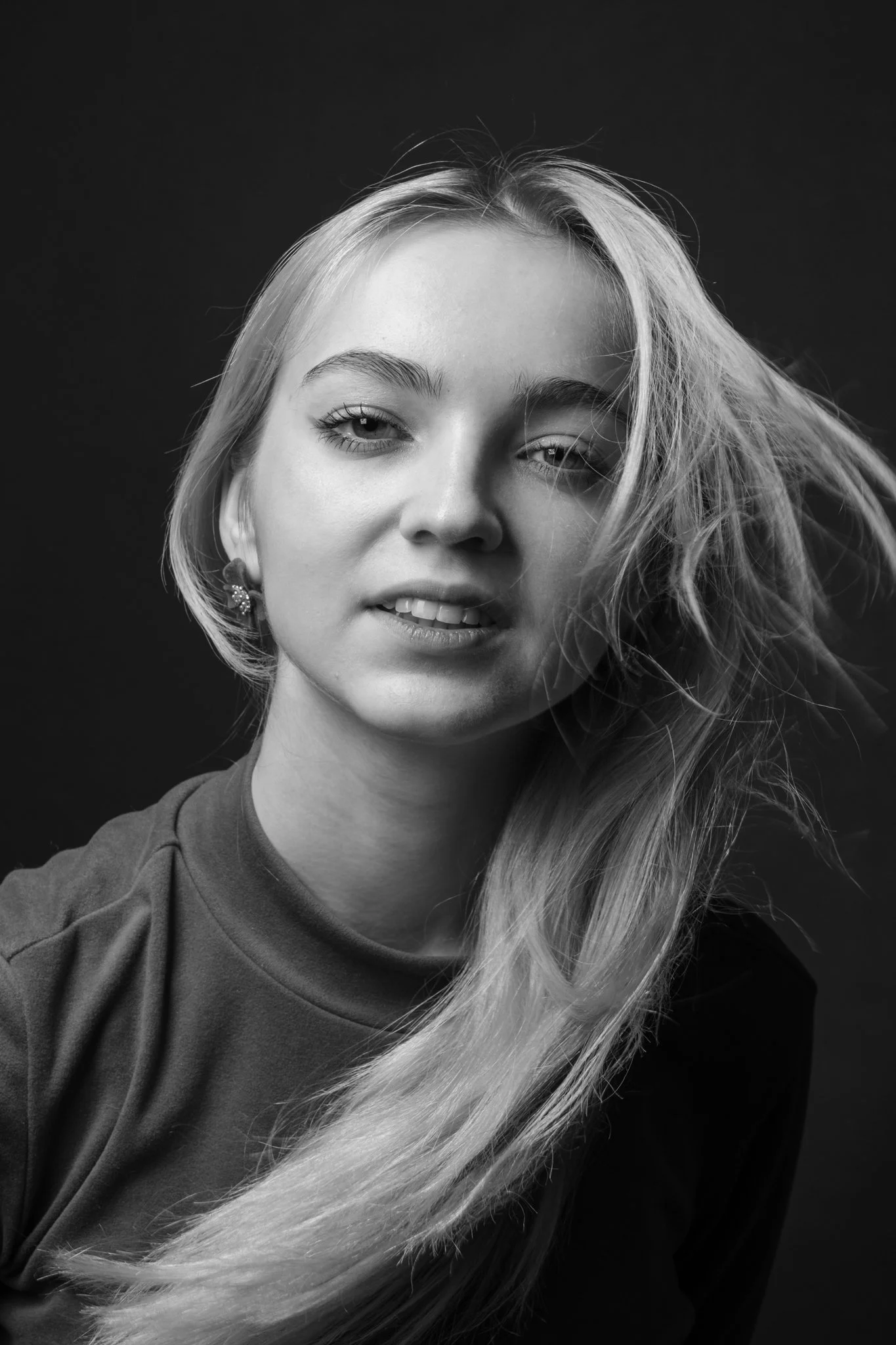 Close-up black and white portrait of a young woman with long, wavy hair, wearing a top with a high neckline and earrings, against a dark background.