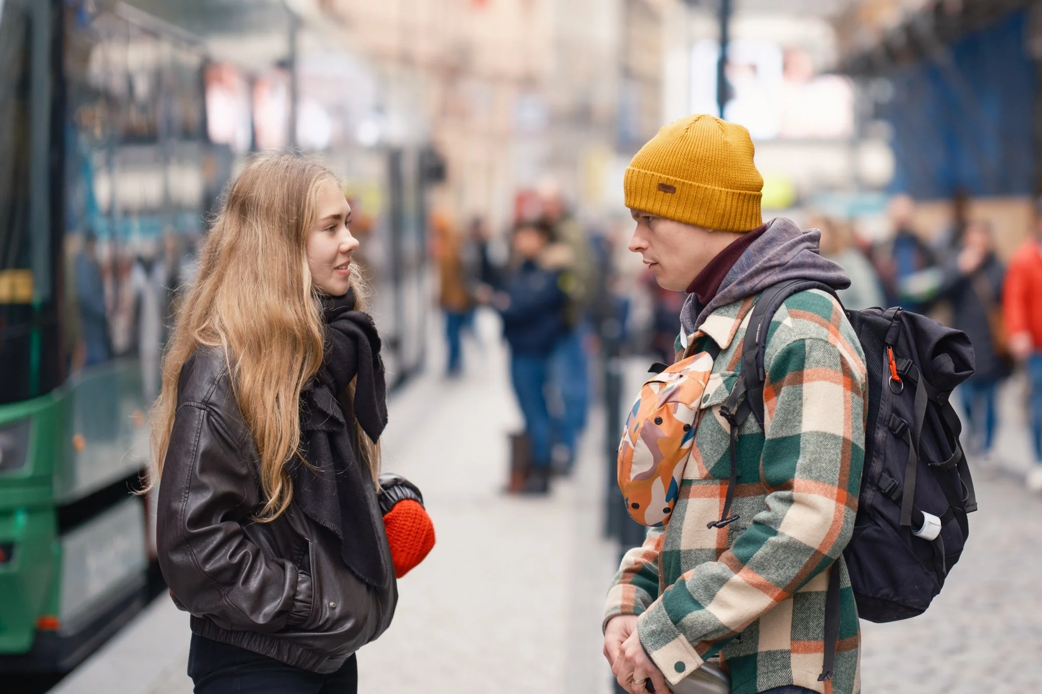 A young man and woman are standing face to face on a busy city street, engaged in conversation. The woman has long blonde hair, is wearing a black leather jacket, and red gloves. The man is wearing a yellow beanie, a plaid jacket, and has a backpack; he appears to be holding a beverage.