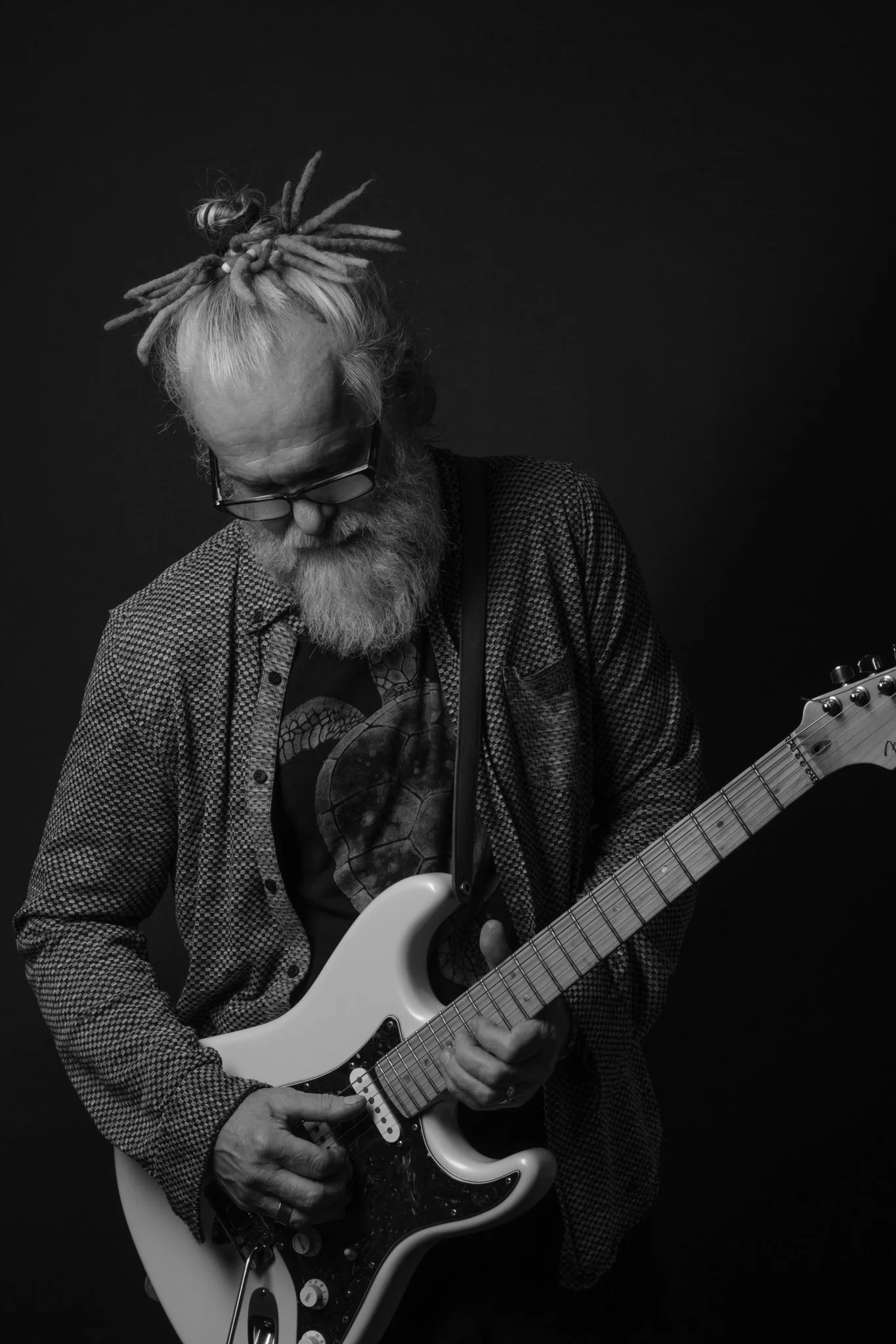 A man with a long beard, glasses, and dreadlocks tied up, wearing a patterned shirt and a graphic t-shirt, playing an electric guitar against a dark background.