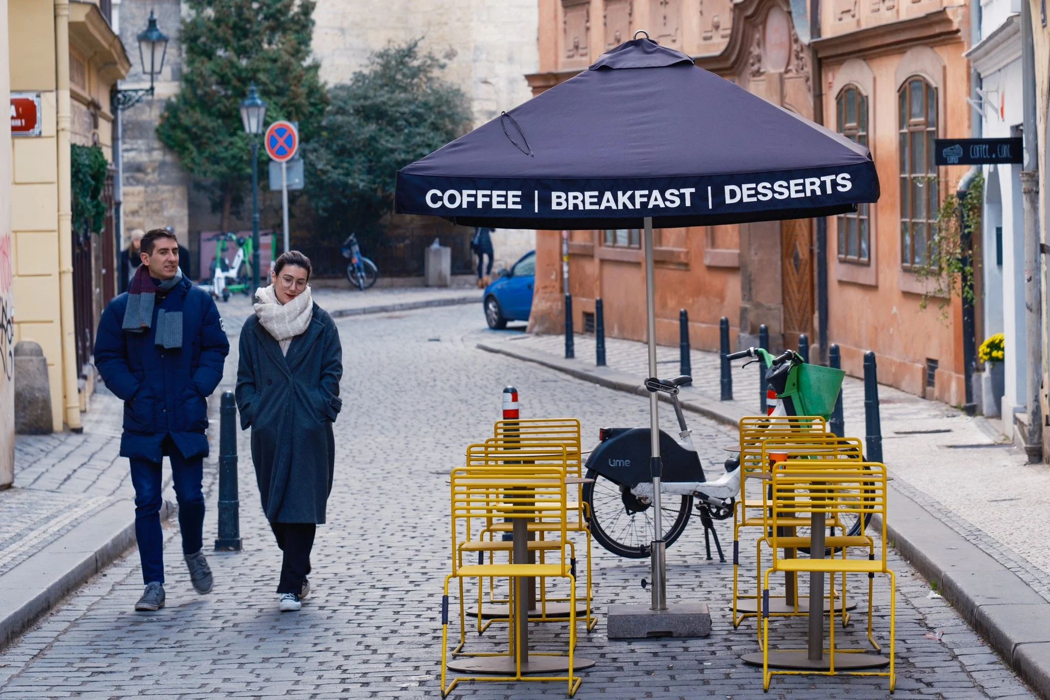 Two people walking on a cobblestone street past an outdoor cafe setup with yellow chairs and a black umbrella that has words advertising coffee, breakfast, and desserts.