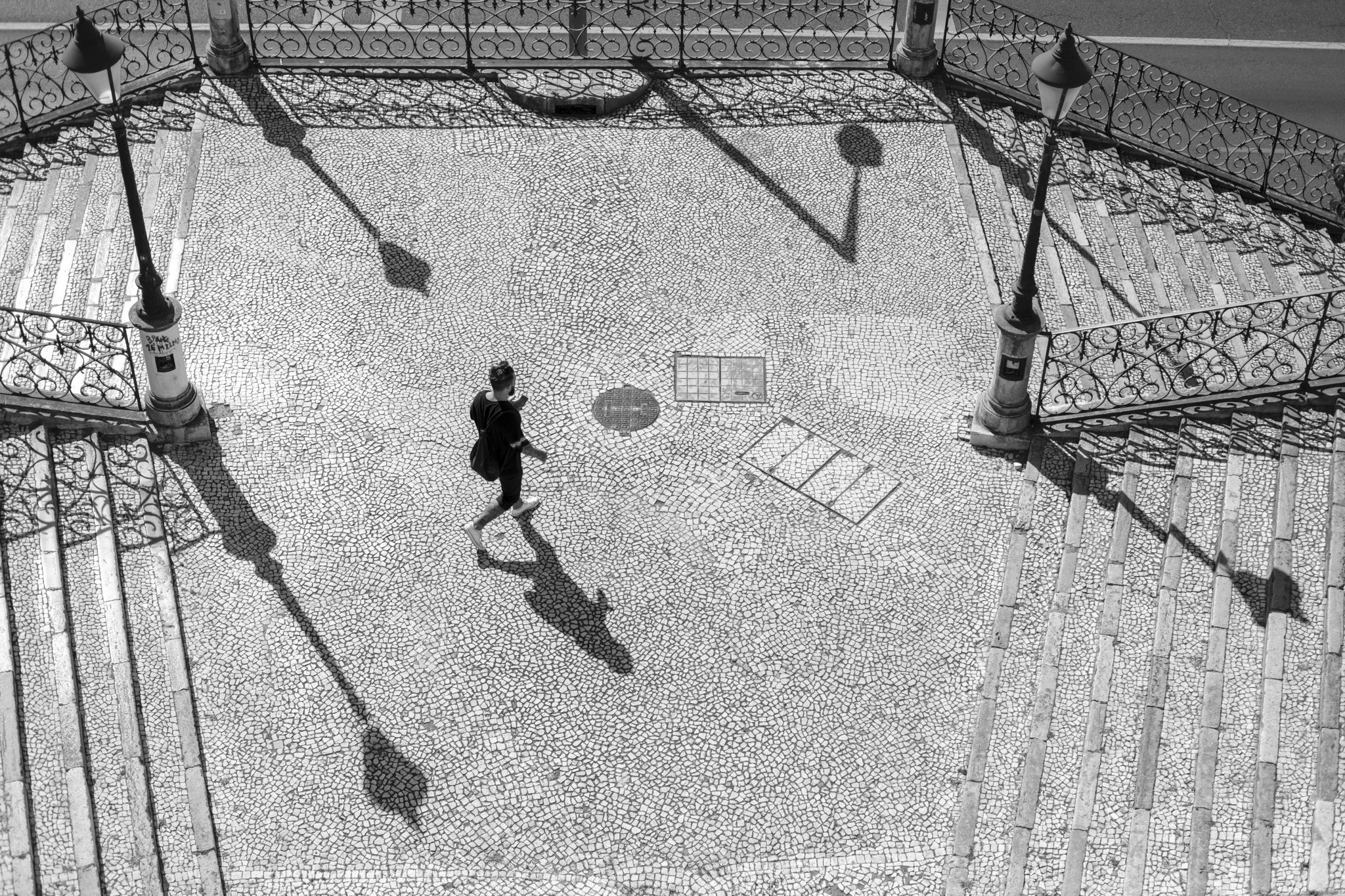 Black and white photo of a person walking on a cobblestone street, with decorative lamp posts casting shadows on the ground, and street railings on the sides.