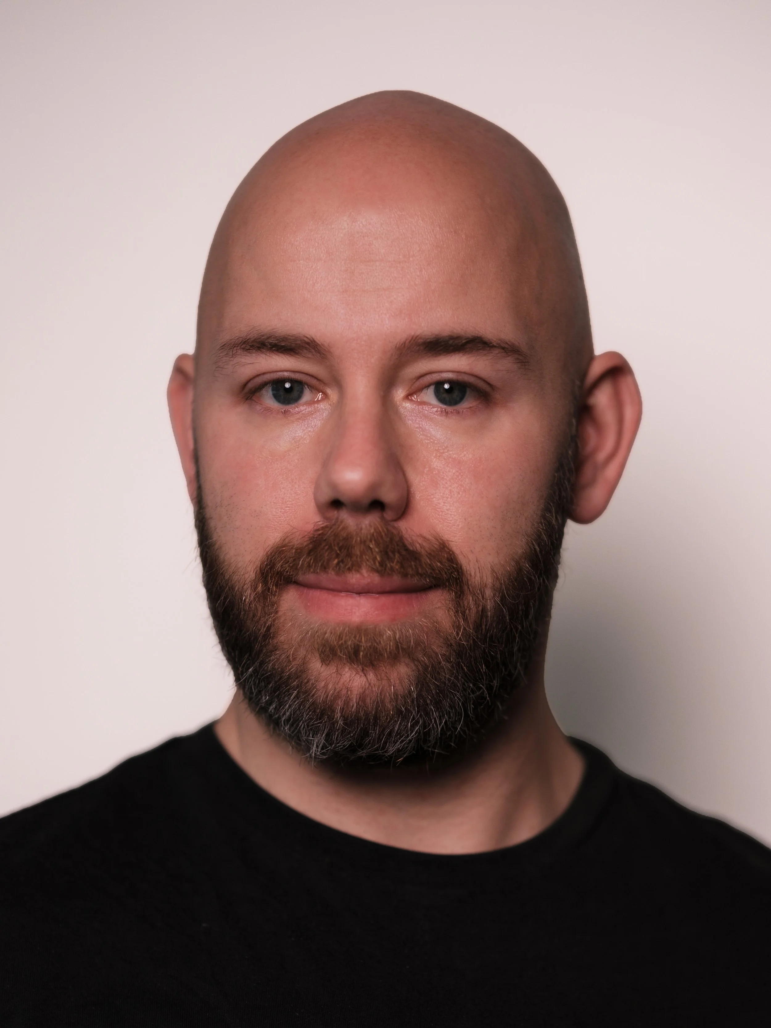Close-up portrait of a bald man with blue eyes, a full beard, and wearing a black shirt, against a plain white background.