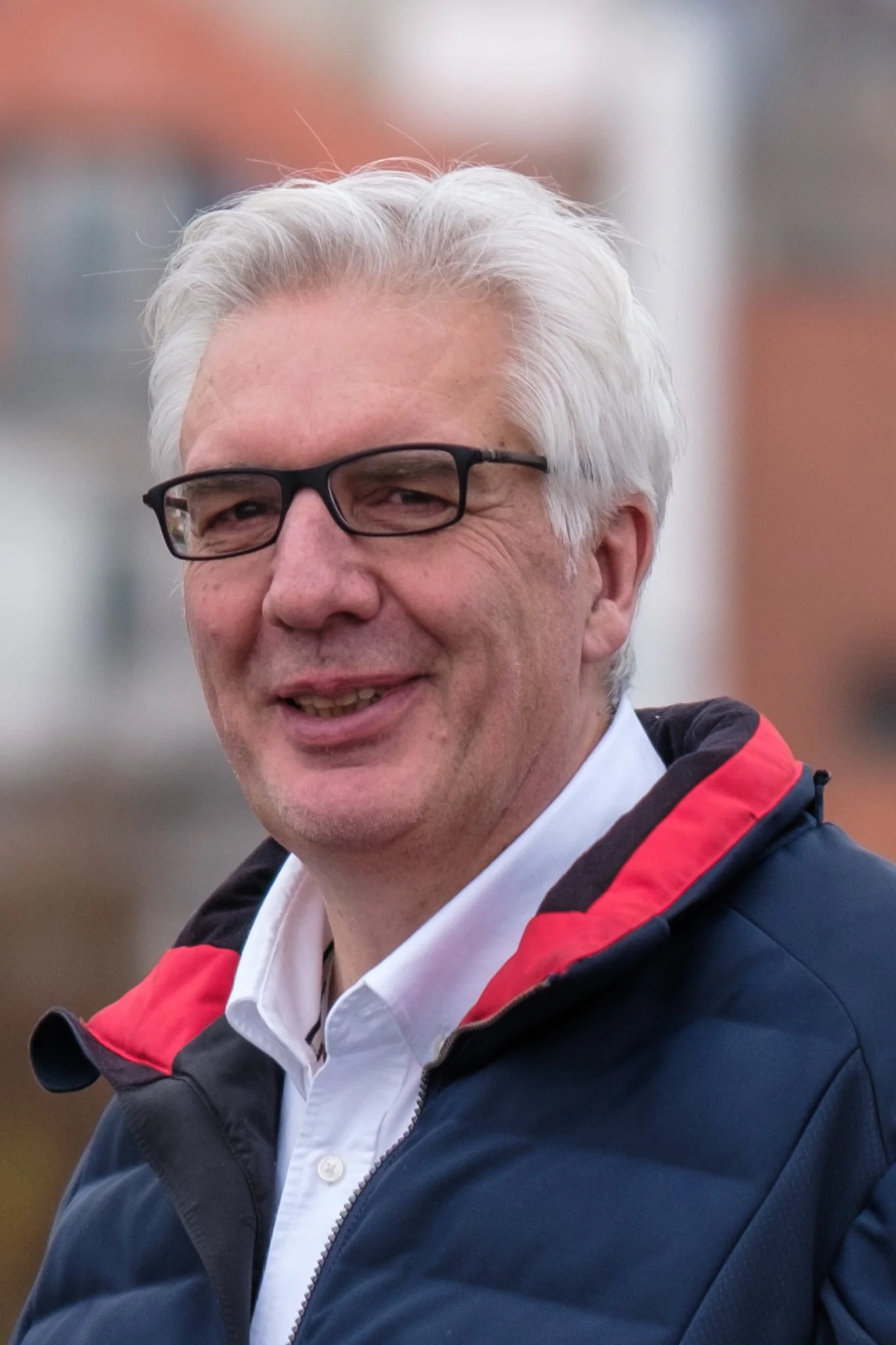 A smiling man with gray hair, wearing black glasses, a white shirt, and a navy blue jacket with red accents, outdoors with blurred buildings in the background.