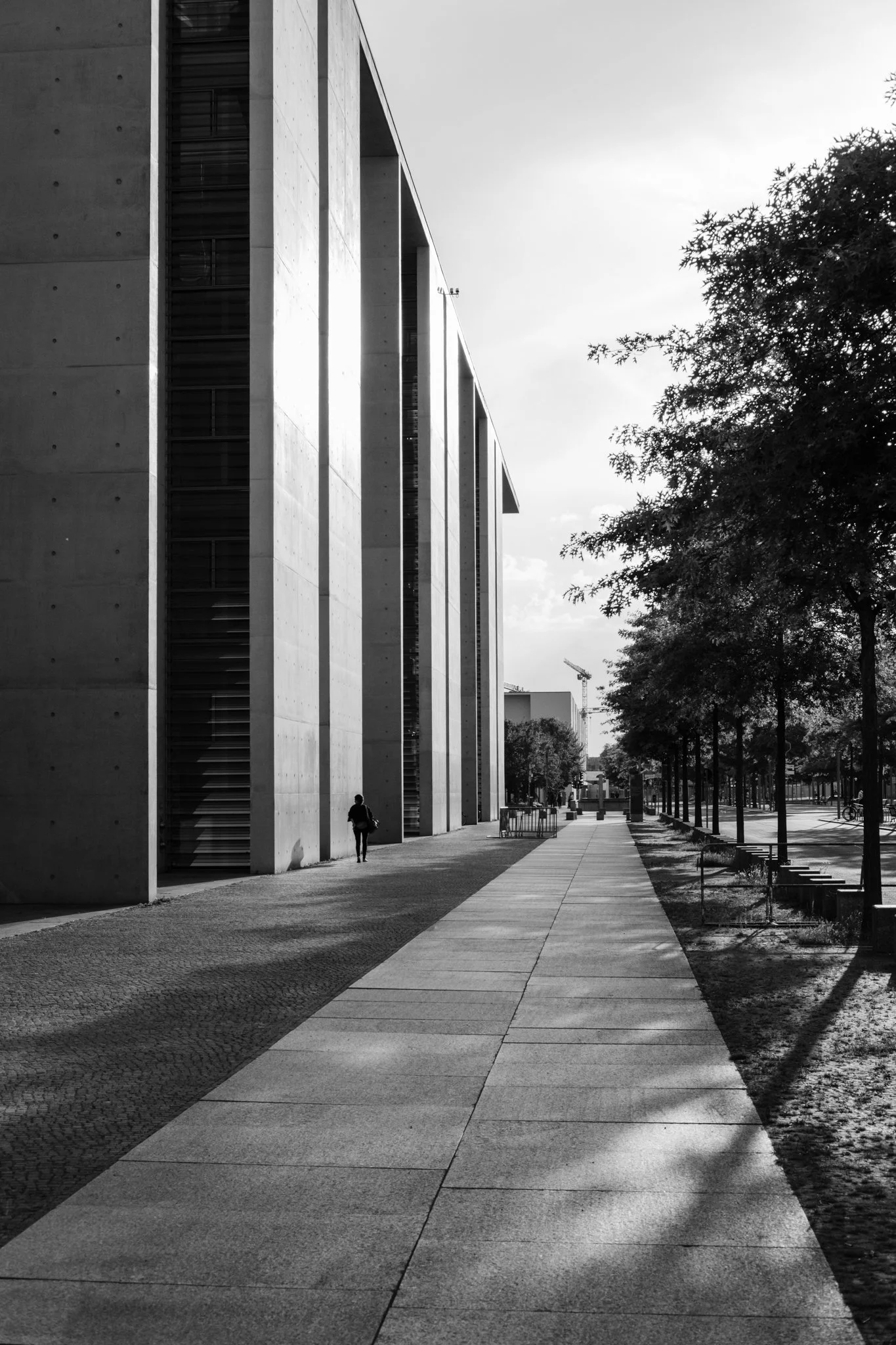 Black and white photograph of a modern building with large concrete walls, a sidewalk, trees on the right, and a person walking along the sidewalk.