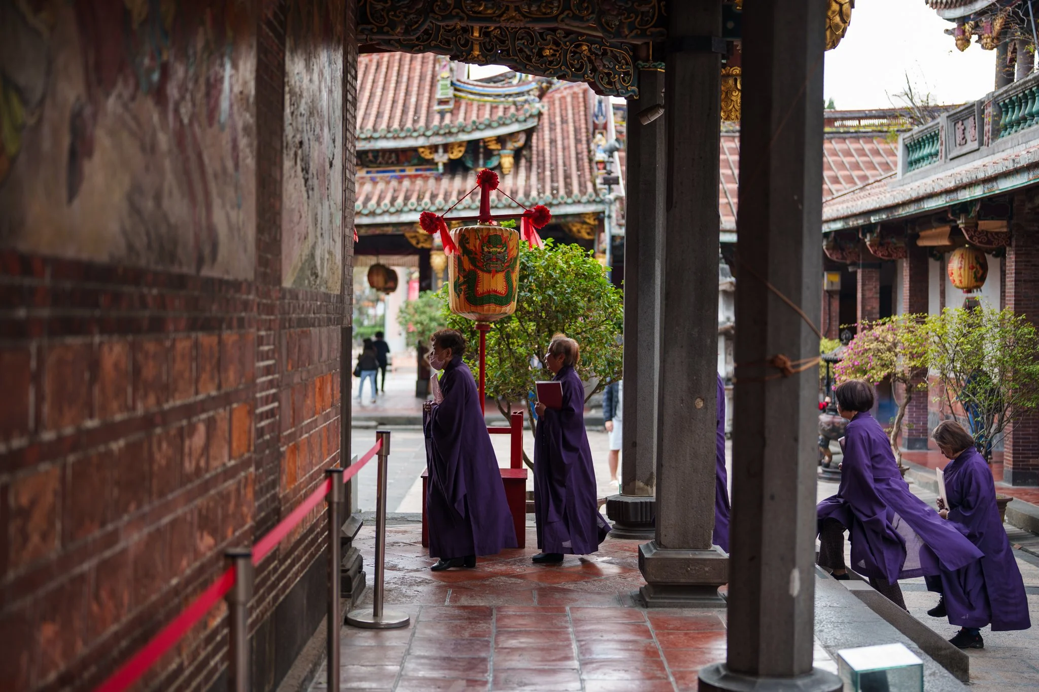 People dressed in purple robes participating in a religious or cultural ceremony inside a traditional Asian temple, with ornate roof designs and hanging lanterns.