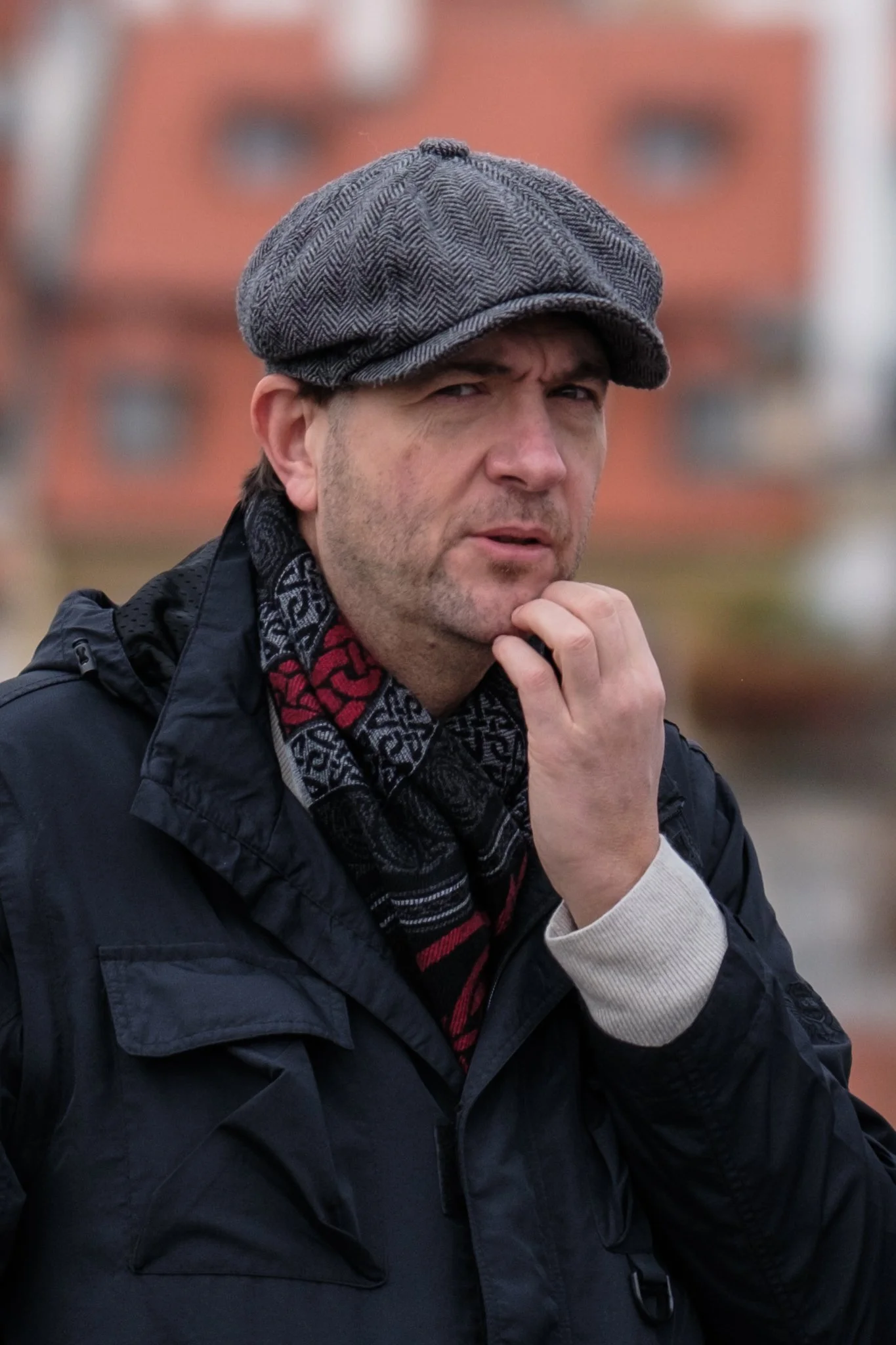 Man wearing a gray checkered flat cap, black jacket, and scarf, standing outdoors with a blurred building background, scratching his chin and furrowing his brow.
