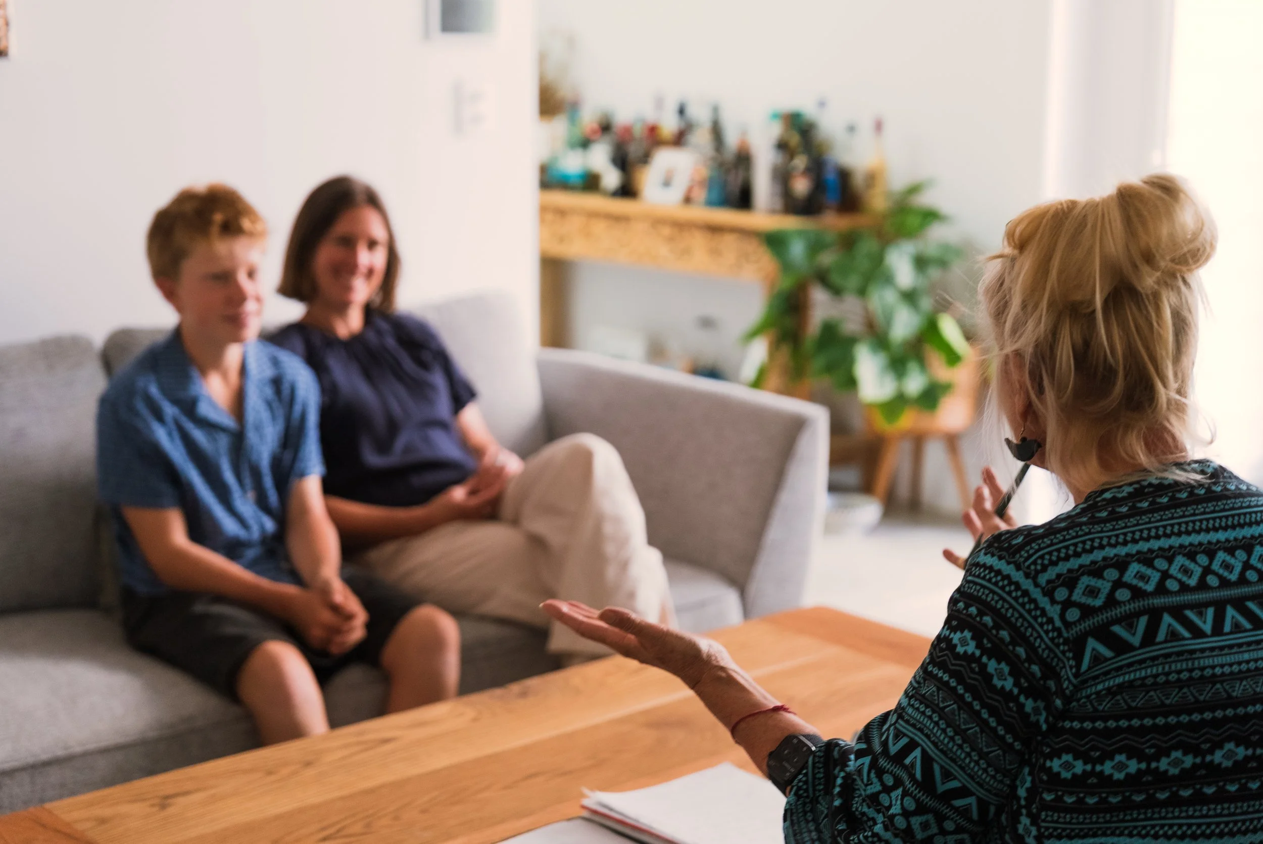 A woman with blonde hair, wearing a patterned sweater, sitting at a wooden table, talking to a boy and a woman sitting on a gray sofa in a living room with plants.