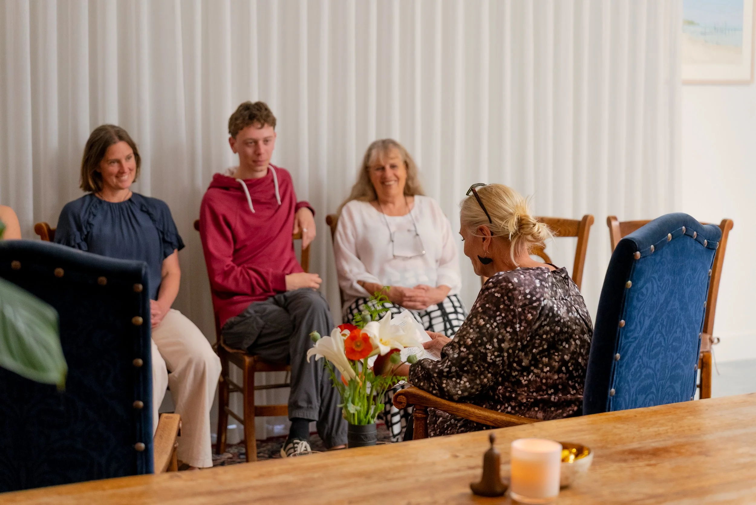 A woman reading a card or letter to a group of four people seated in a room with white curtains. The group appears engaged and happy, with a vase of flowers on the table and candles in the foreground.