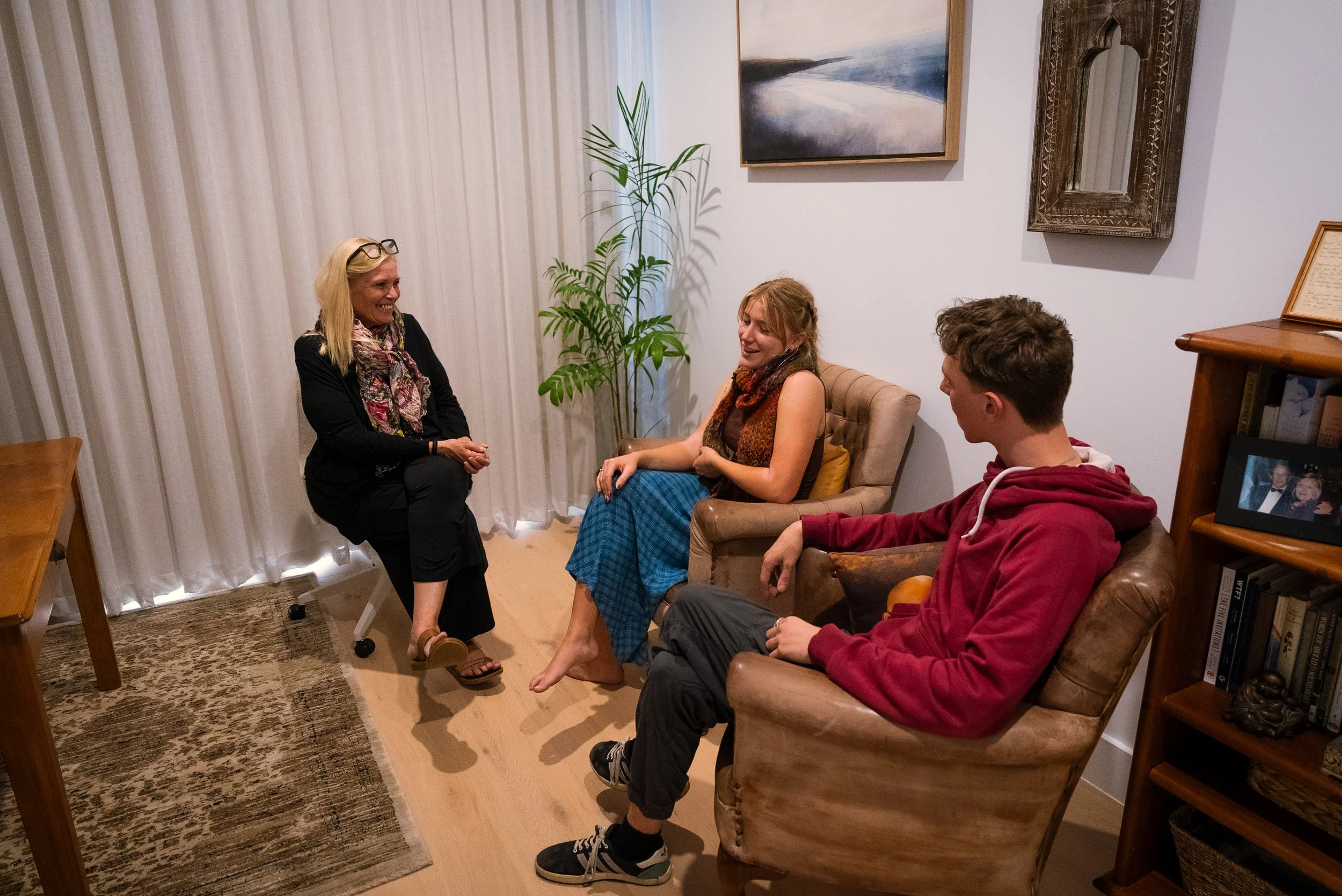 Three people, a woman and two teenagers, sit and stand in a living room, engaging in conversation and smiling. The woman is sitting on a chair, the girl is on a beige armchair, and the boy is in a brown leather armchair. The Self Sanctuary