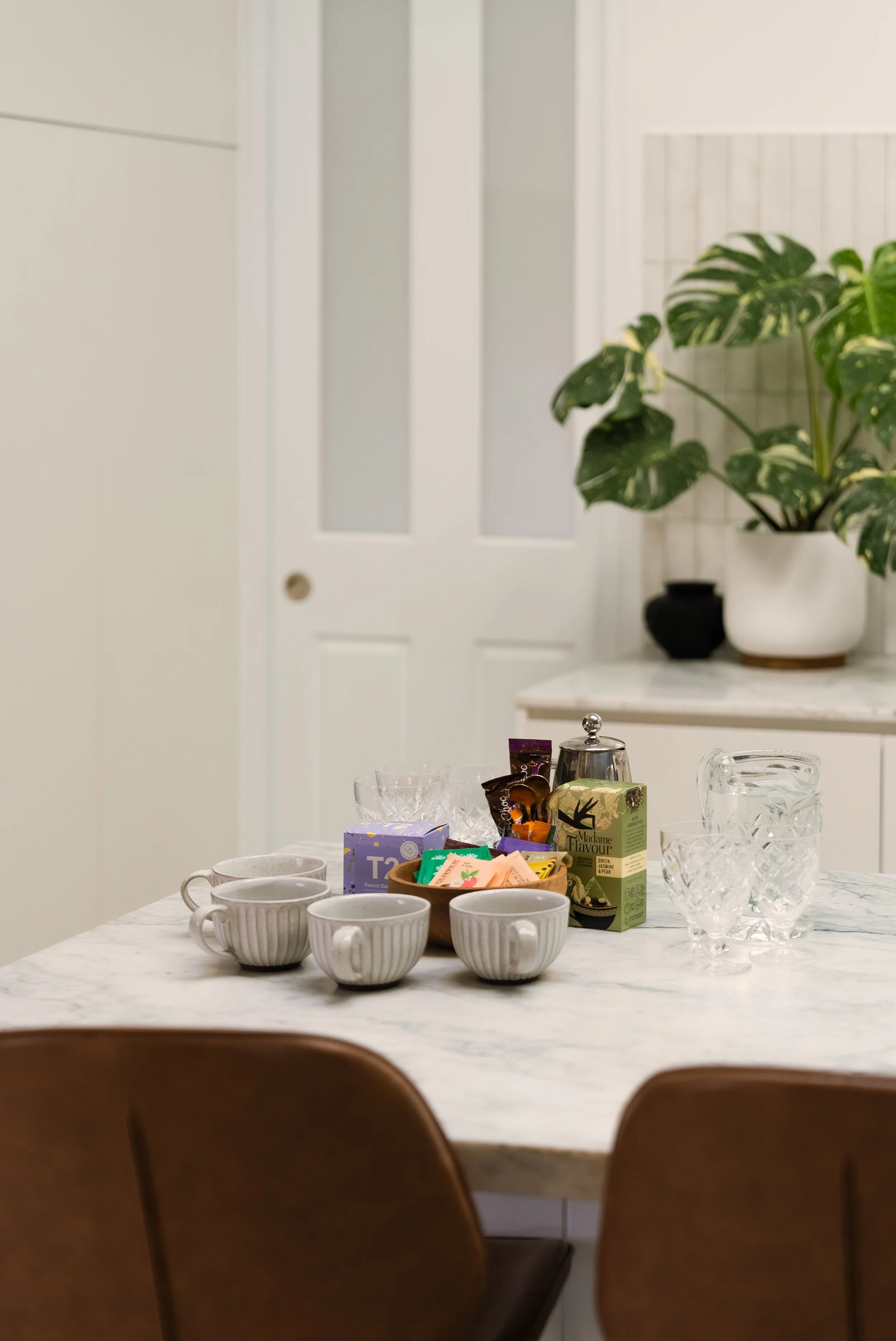 A marble dining table with four white ceramic cups, a bowl of tea bags, two clear glass cups, and a small metal teapot. In the background, there's a large houseplant in a white pot on a white marble surface, and a white door in a decorated interior.