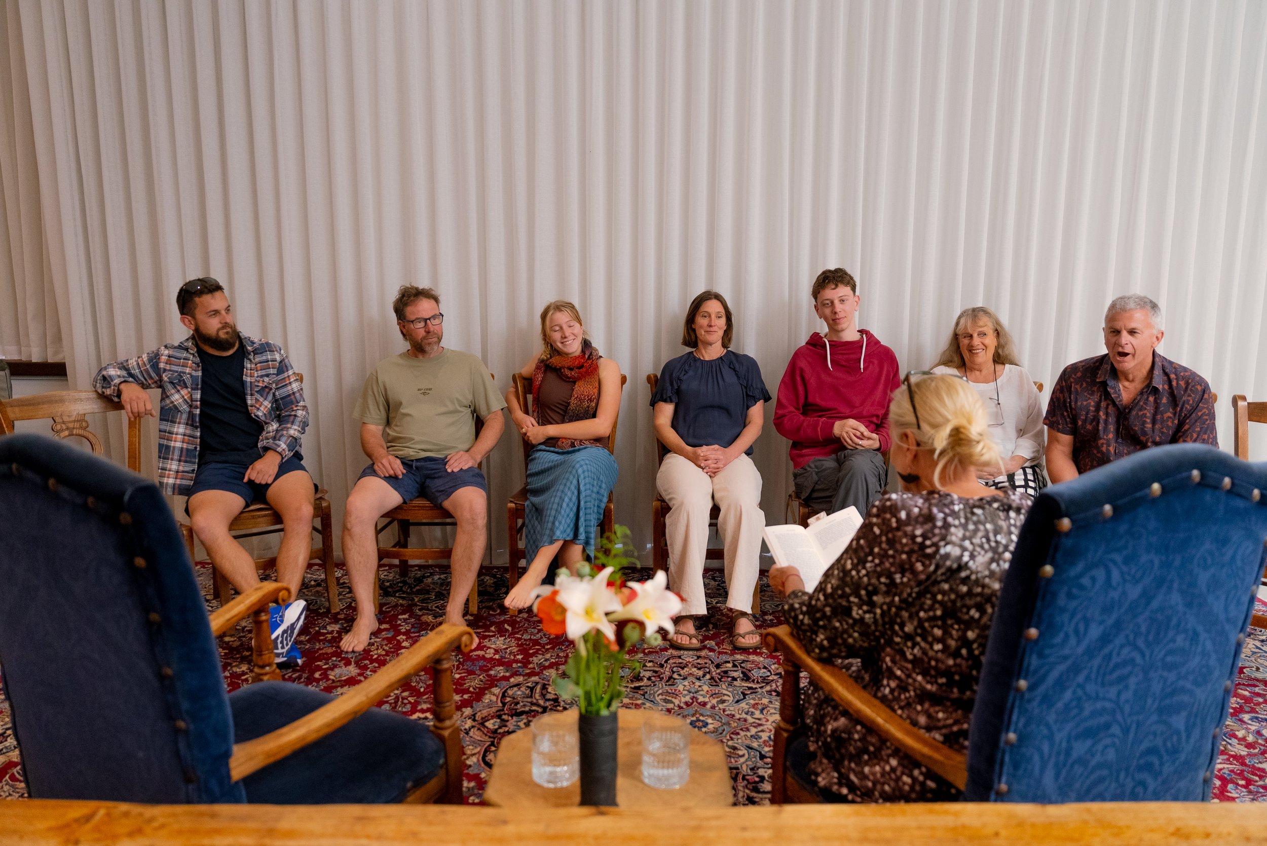 A woman reading a card or letter to a group of four people seated in a room with white curtains. The group appears engaged and happy, with a vase of flowers on the table and candles in the foreground.