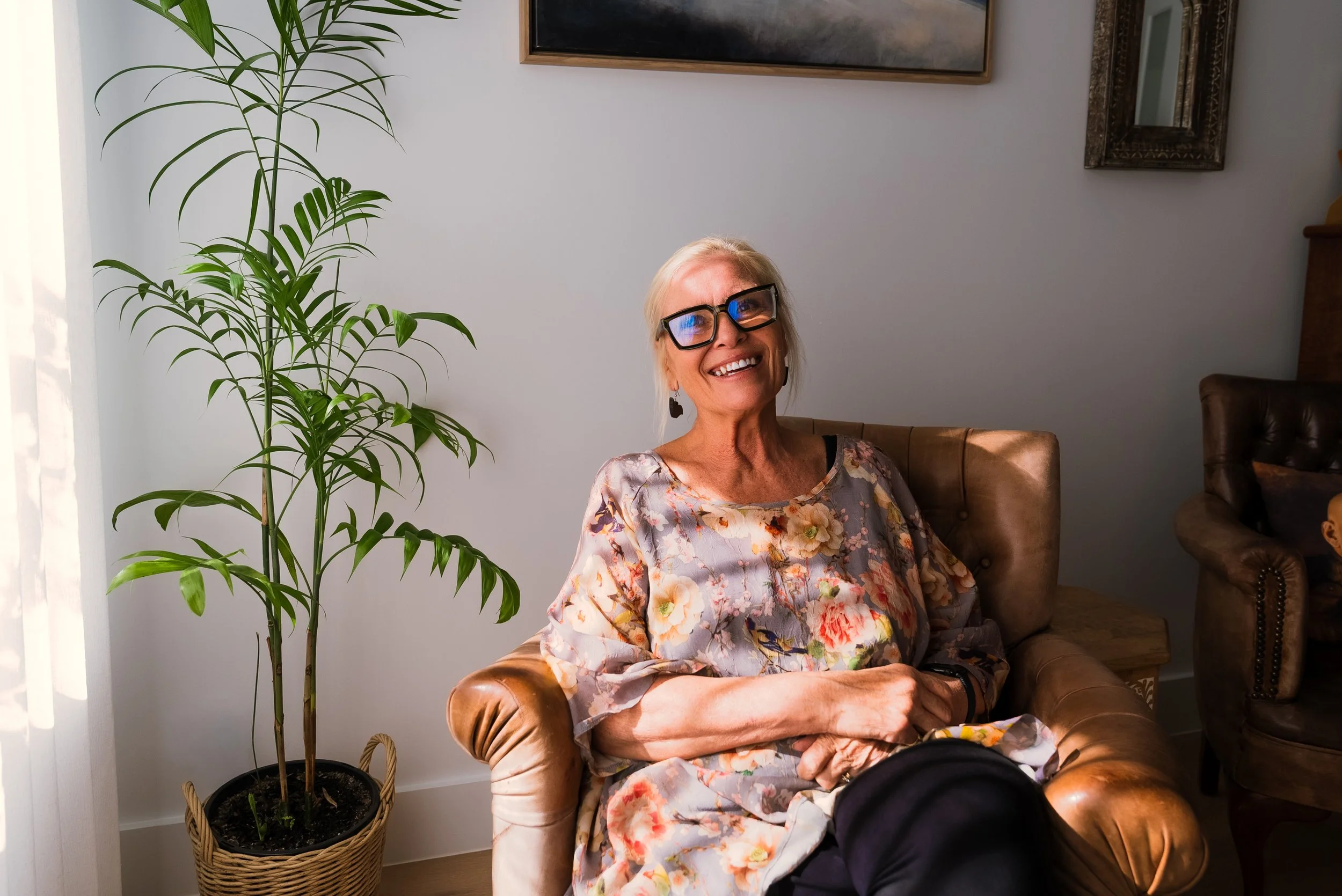 An experienced woman with glasses and tied blonde hair sitting on a leather armchair, smiling, in a well-lit room. There is a tall green plant in a woven basket to her left and framed artwork on the wall behind her. The Self Santuary