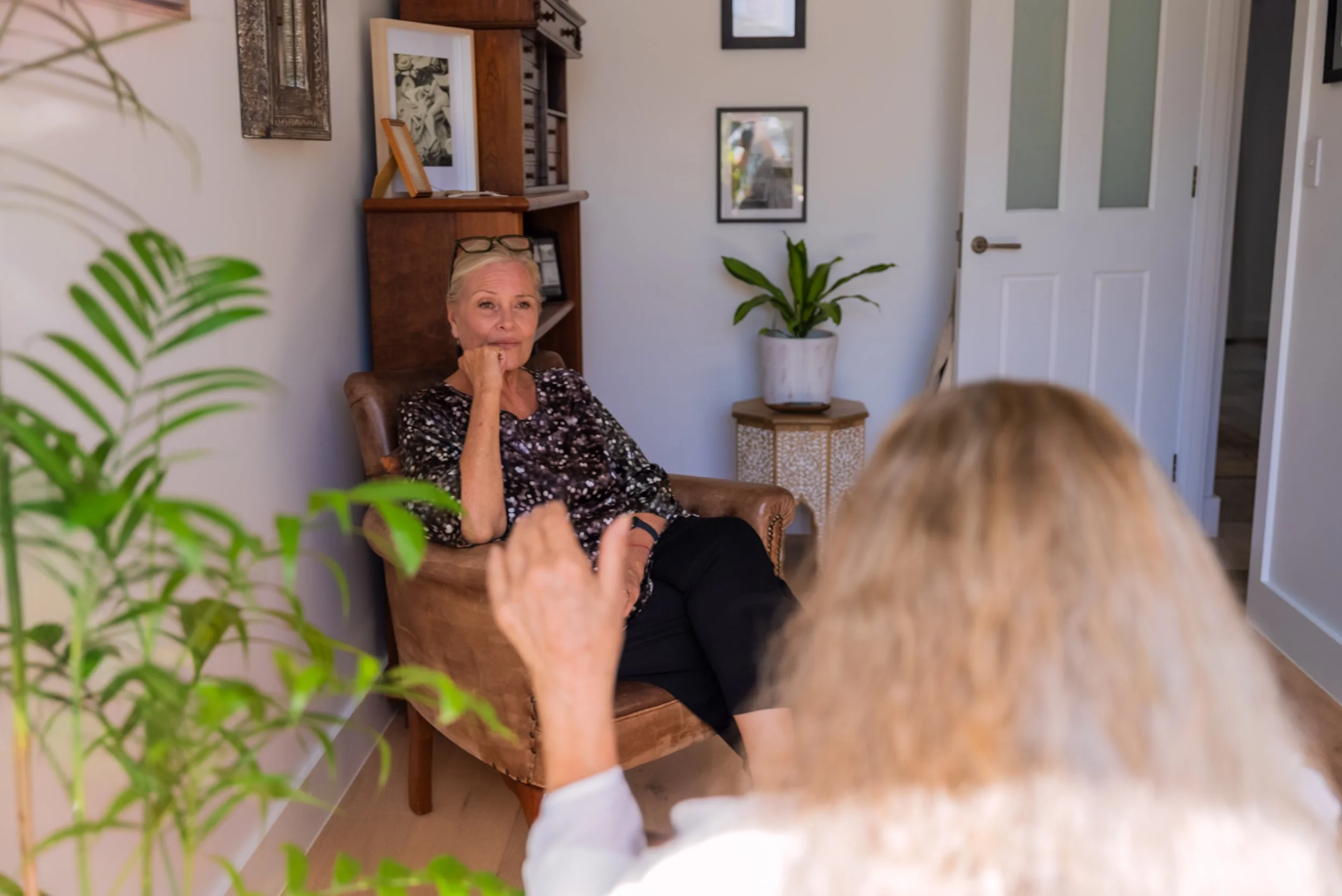 A woman sits in a brown armchair during a conversation with a person whose back is towards the camera. The woman appears thoughtful as she listens or speaks. The setting is a cozy room with framed pictures on the wall, a large potted plant on a side table, and a wooden bookshelf filled with books and decorative items.