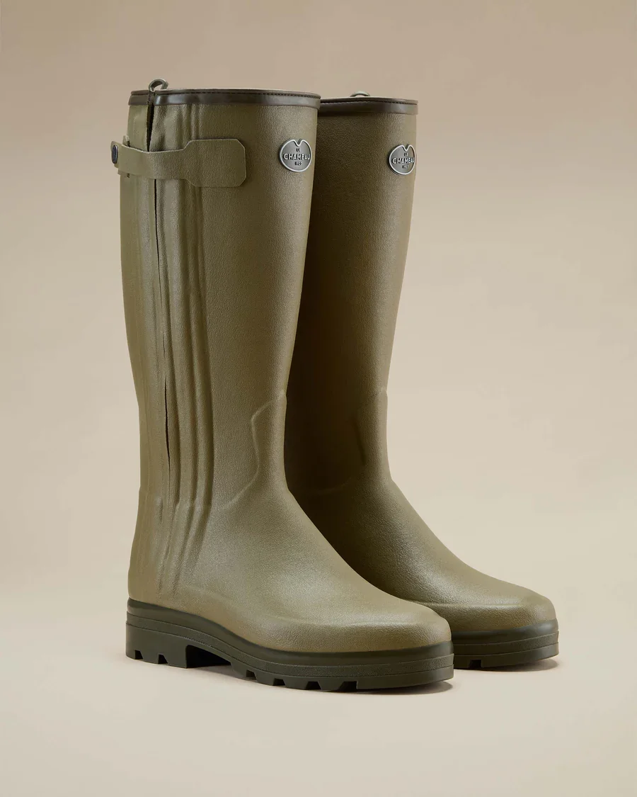 Pair of olive green rubber rain boots with black soles and metallic emblem logos on the sides, standing upright on a beige background.