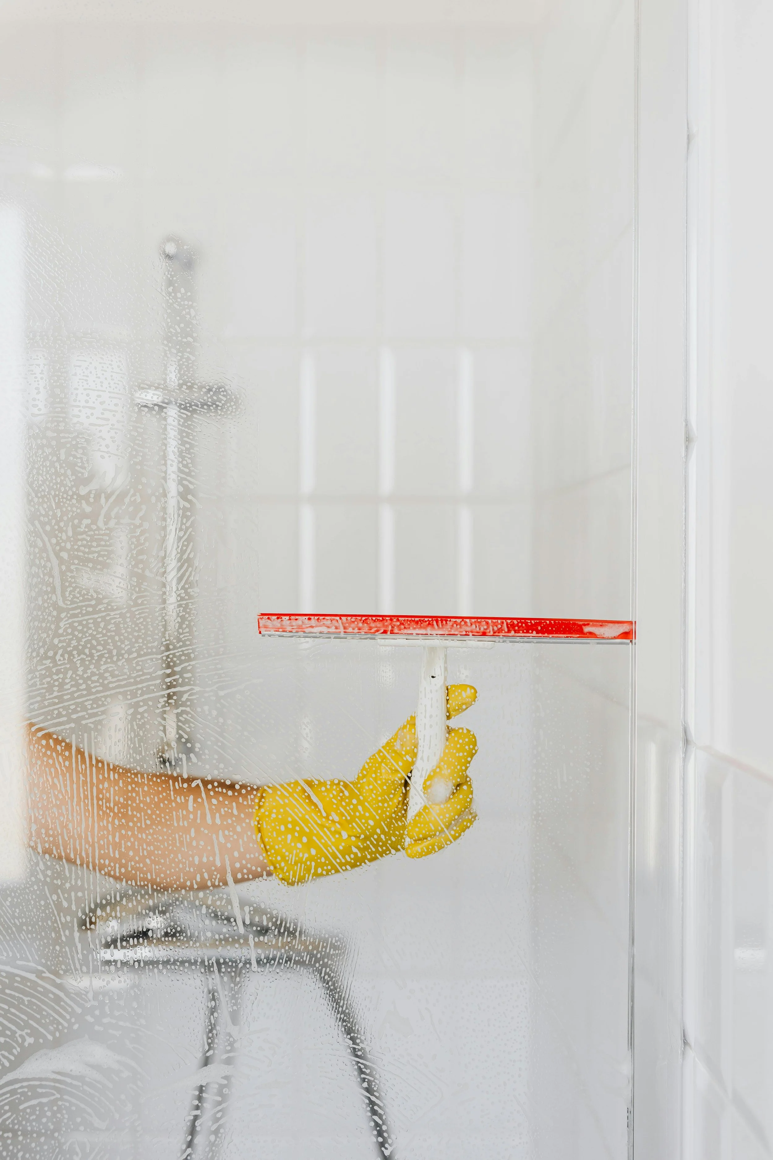Person cleaning a glass shower door with a squeegee, wearing yellow rubber gloves.