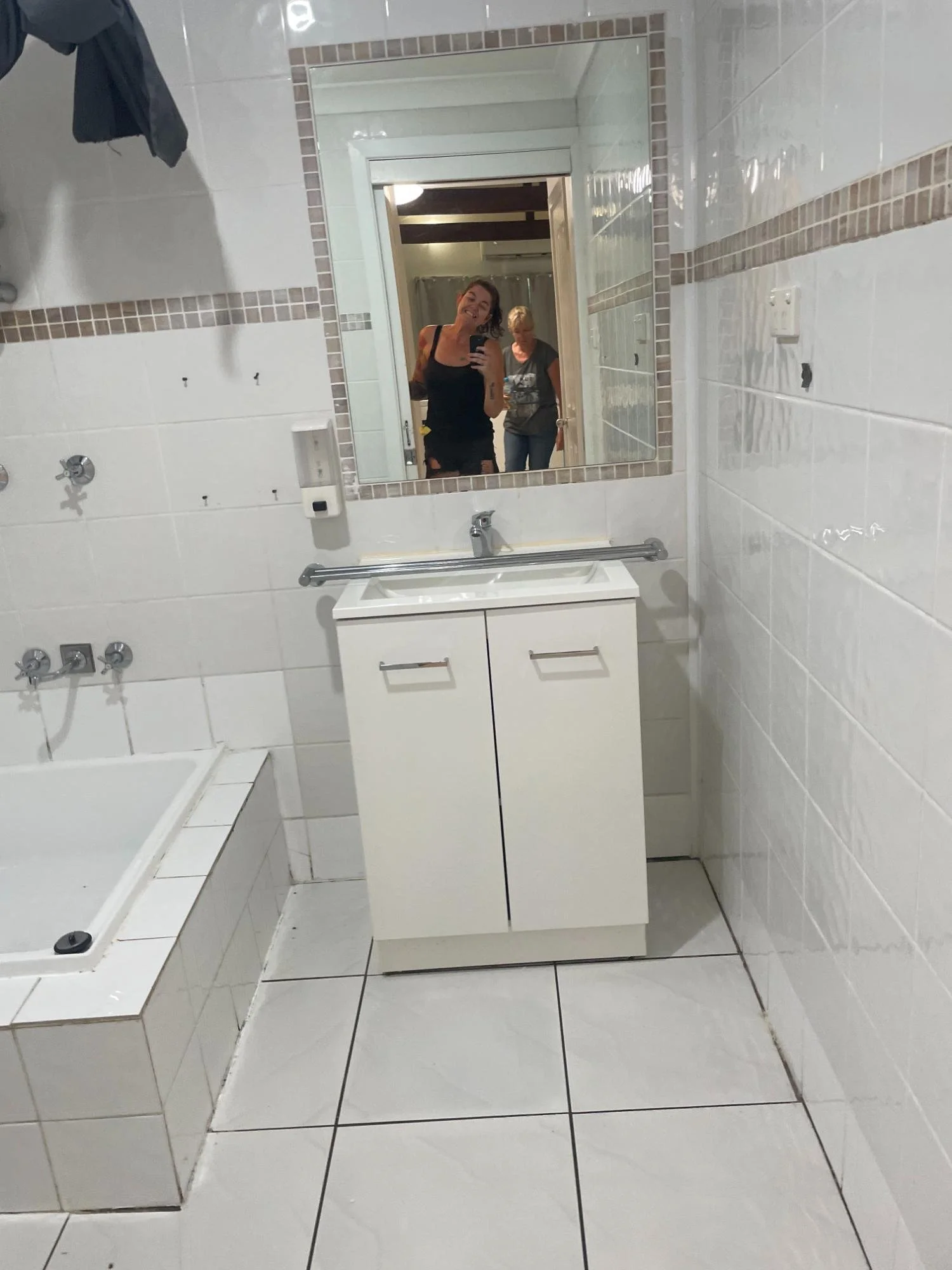 A bathroom with a white cabinet and sink, large mirror, and a bathtub. Two women are reflected in the mirror, one smiling and taking a photo, and the other looking down.