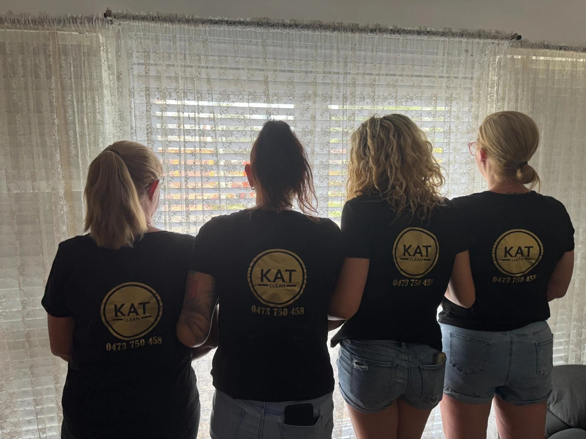Four women standing indoors, facing a window with lace curtains. They are wearing matching black t-shirts with a yellow logo and a phone number on the back, and denim shorts or jeans.