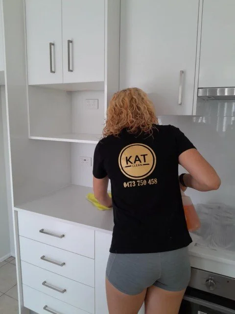 Blonde woman wearing black t-shirt with Kat Clean logo displayed on back, cleaning kitchen bench.