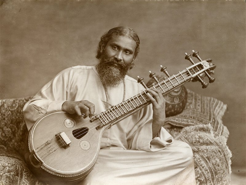 Sepia portret van Hazrat Inayat Khan, die een ketting met een hartvormige hanger draagt, oftewel het Soefisme symbool. Hij zit op een sofa met sierkussens en speelt op een sitar.