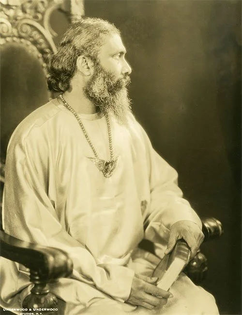 Portret van Hazrat Inayat Khan, zittend op een ornamentale stoel. Hij draagt een satijnen gewaad en een ketting met het soefisme symbool. Hij houdt een boek in zijn rechterhand.