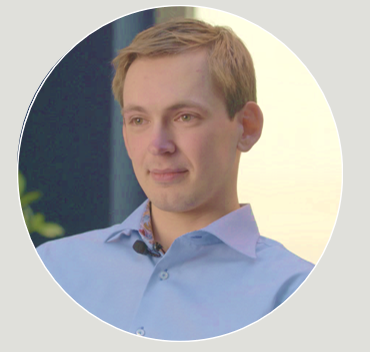 A young man with light brown hair and fair skin, wearing a light blue collared shirt, sitting indoors in a professional or casual setting.