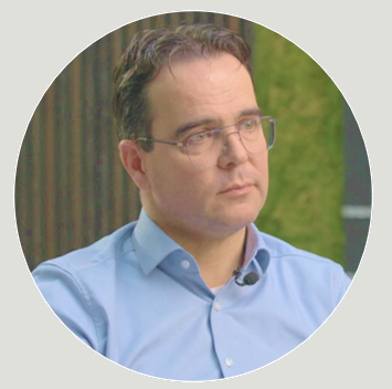 A man with glasses and dark hair wearing a light blue button-down shirt outdoors.