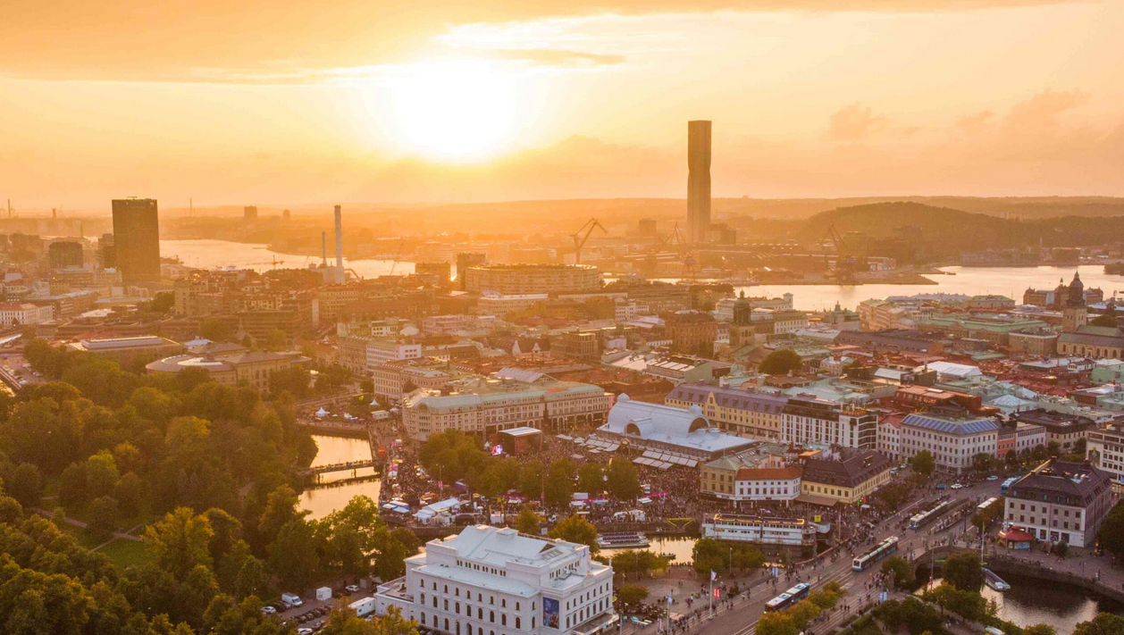 A cityscape at sunset with a river, bridge, and tall buildings in the background, and a park with trees and water in the foreground.
