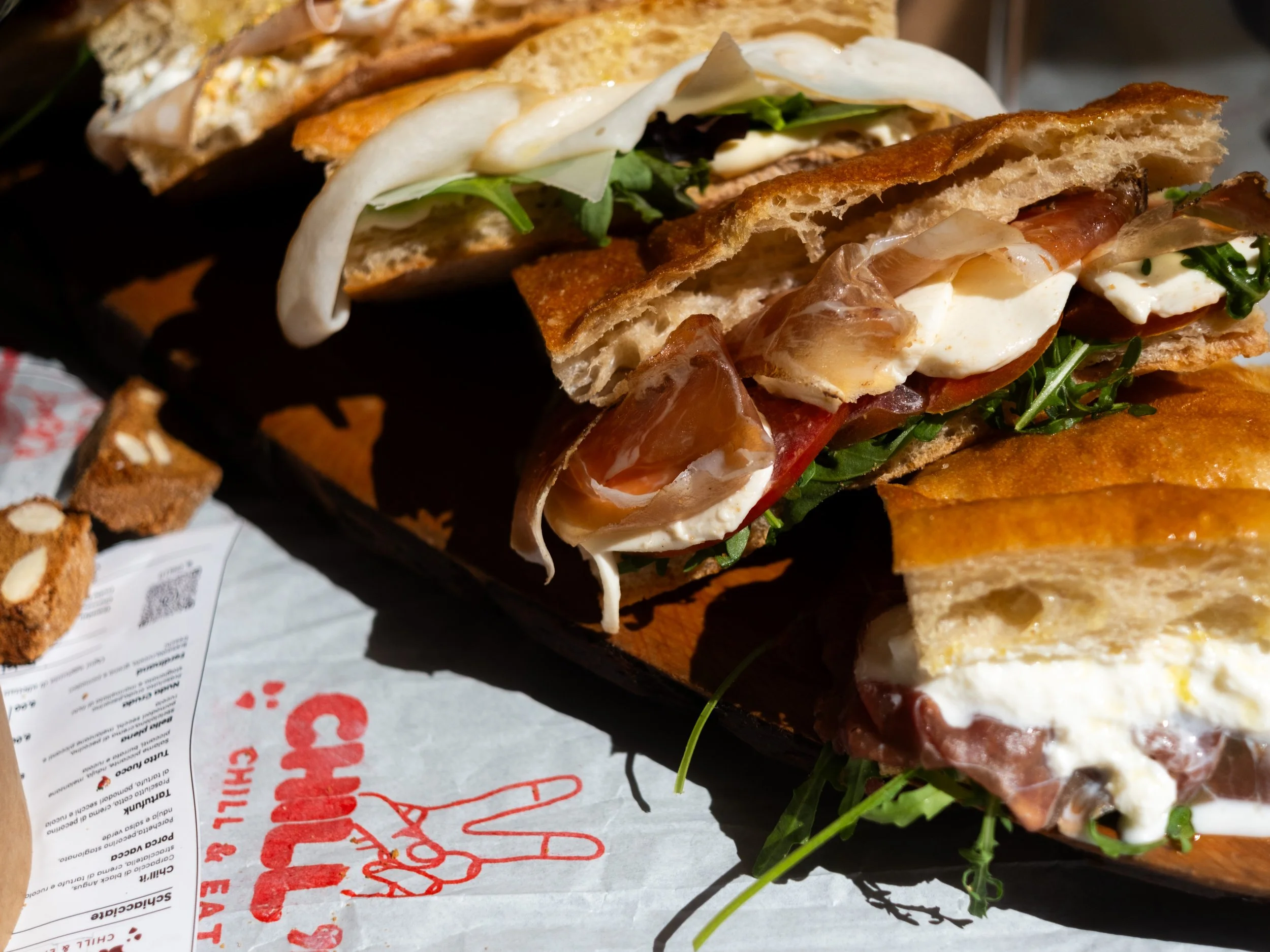 Close-up of a crispy Italian sandwich with layers of cured meats, cheese, fresh greens, and grilled bread, placed on a black tray with restaurant branding.