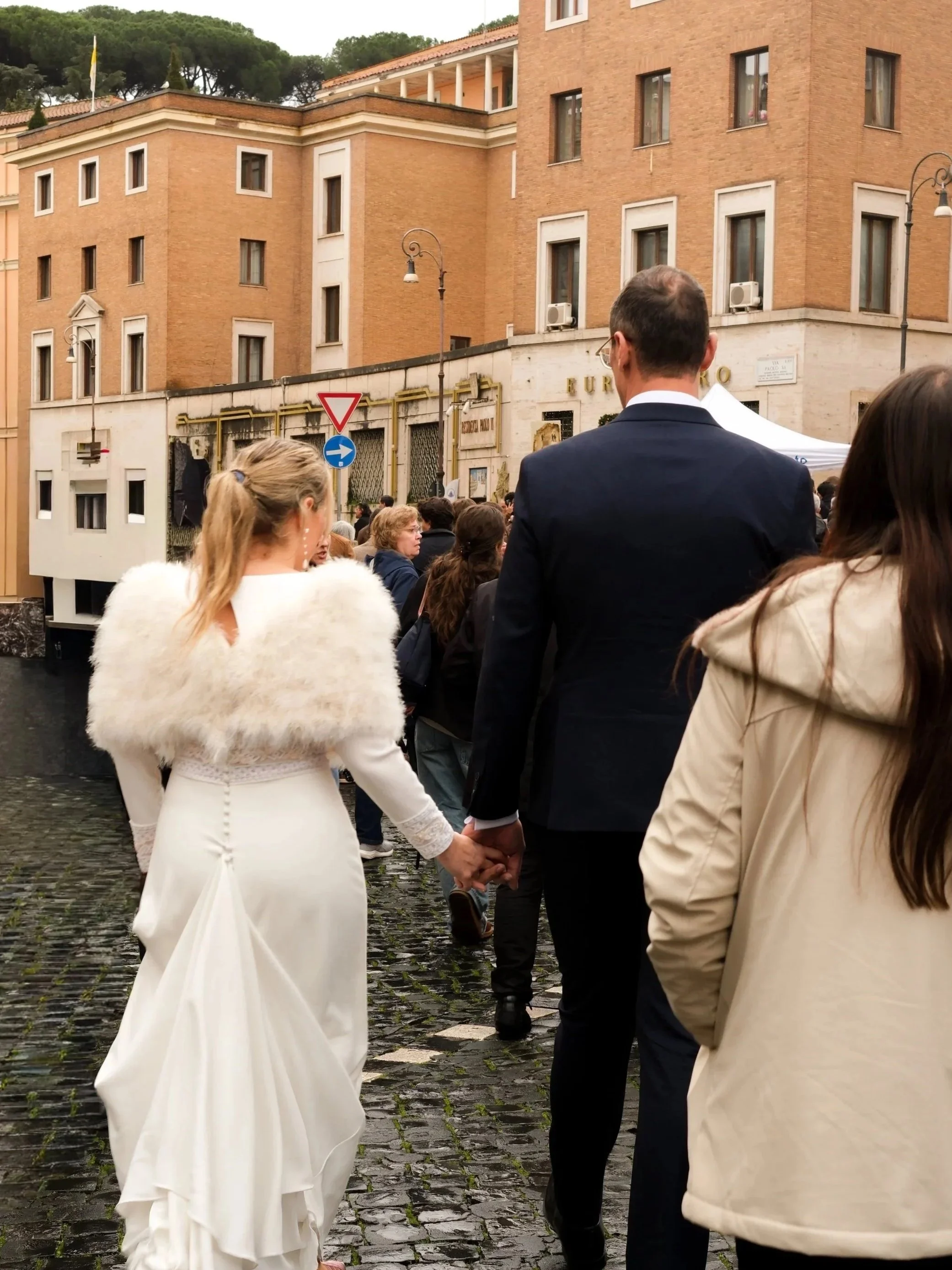 Wedding couple holding hands as they walk through a crowd on cobblestone street in Rome in front of a beige brick building.