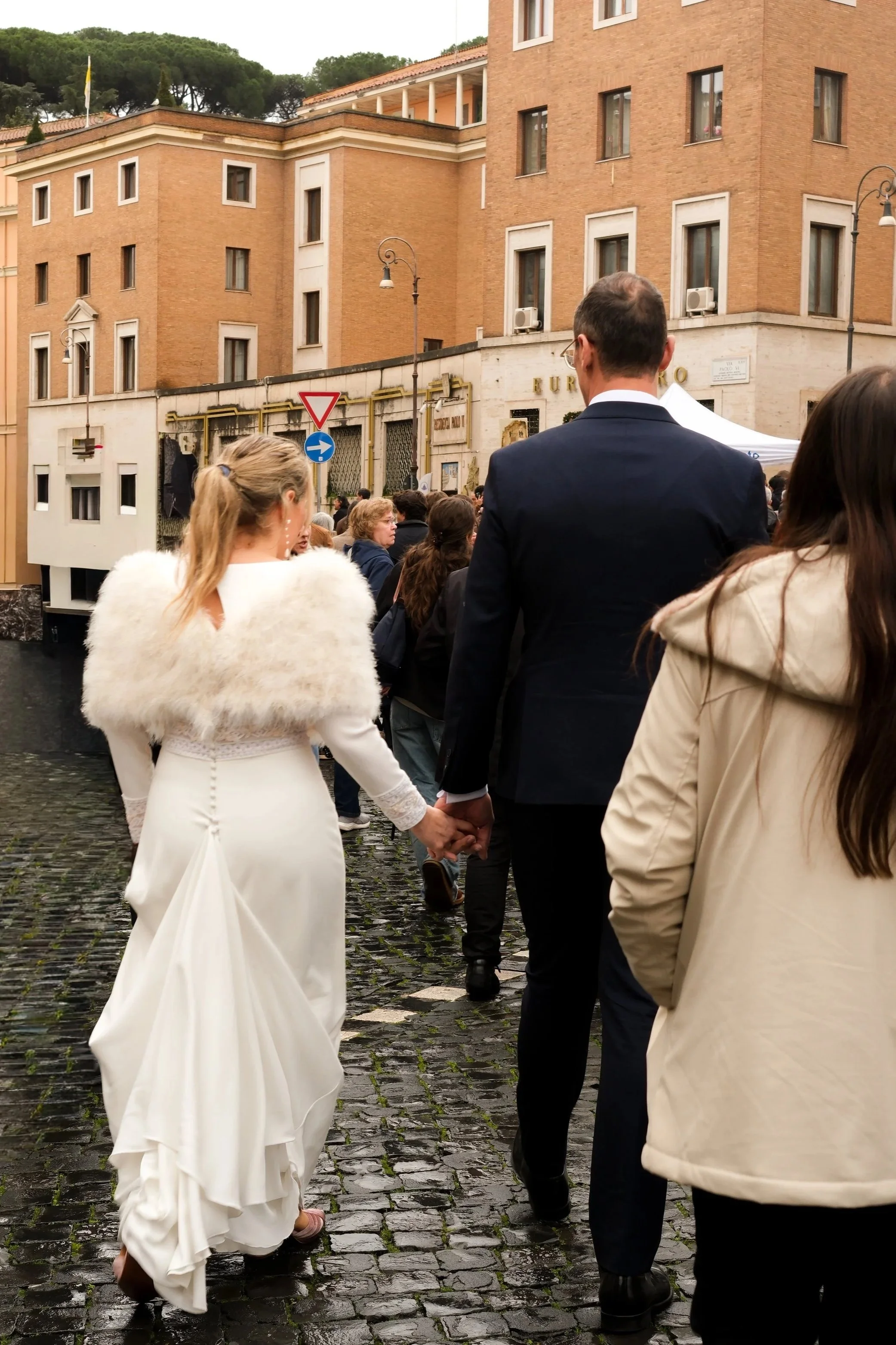 A bride and groom holding hands amidst a crowd of people outdoors in Rome on cobblestone streets, with old European-style buildings in the background.