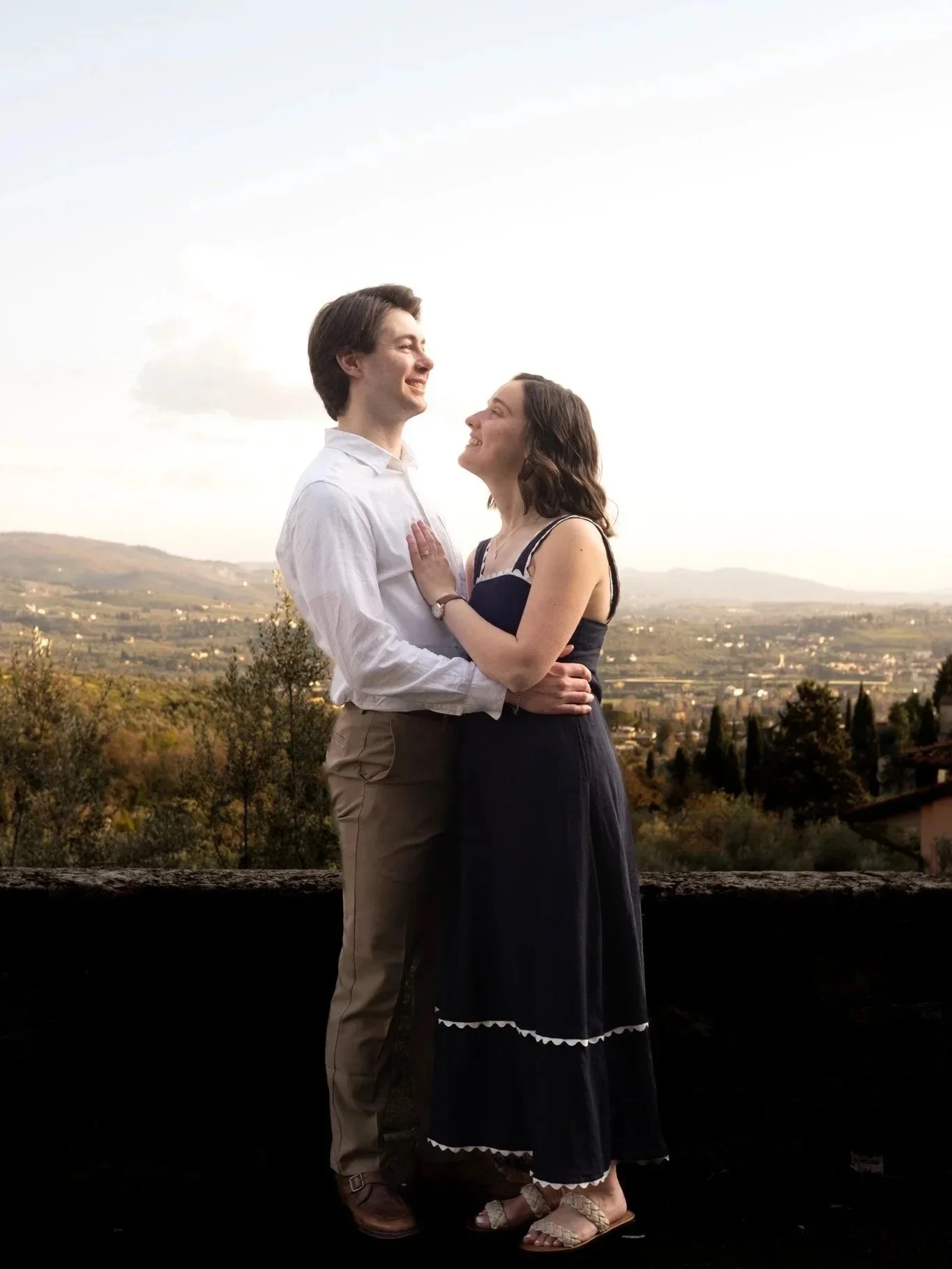 A couple standing outdoors, smiling and looking at each other, with scenic tuscan hills in the background.