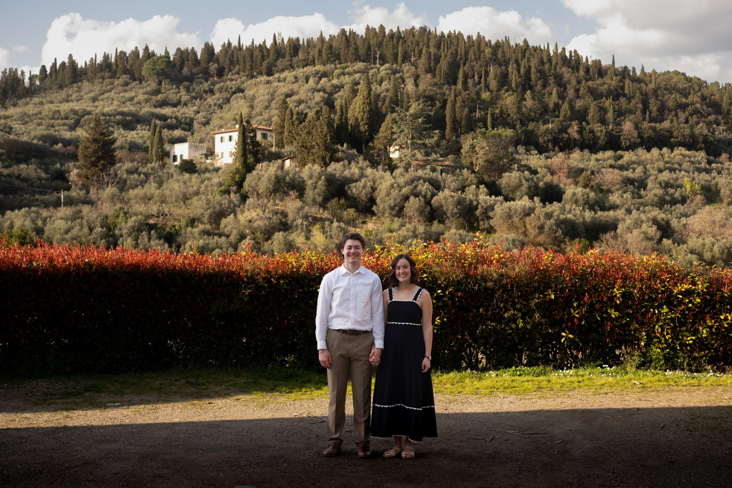 A young couple standing hand in hand outdoors, with a hillside covered in trees and houses in the background, and a red bush in the foreground near Florence, Tuscany, Italy.