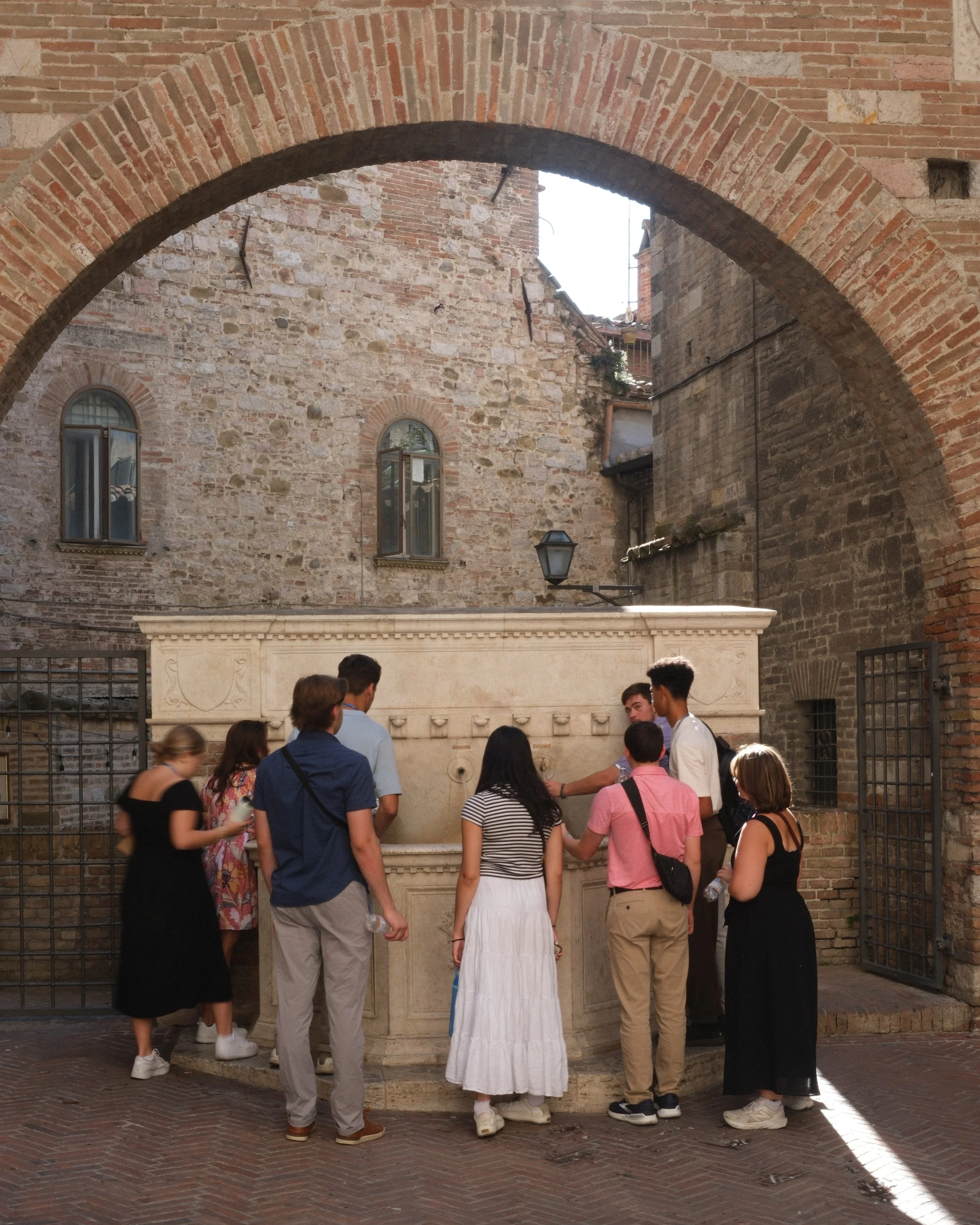 Group of nine young people on a tour in Perugia, Umbria, Italy gathered around a stone fountain in a brick courtyard with an arched brick entryway, stone walls, and small windows.