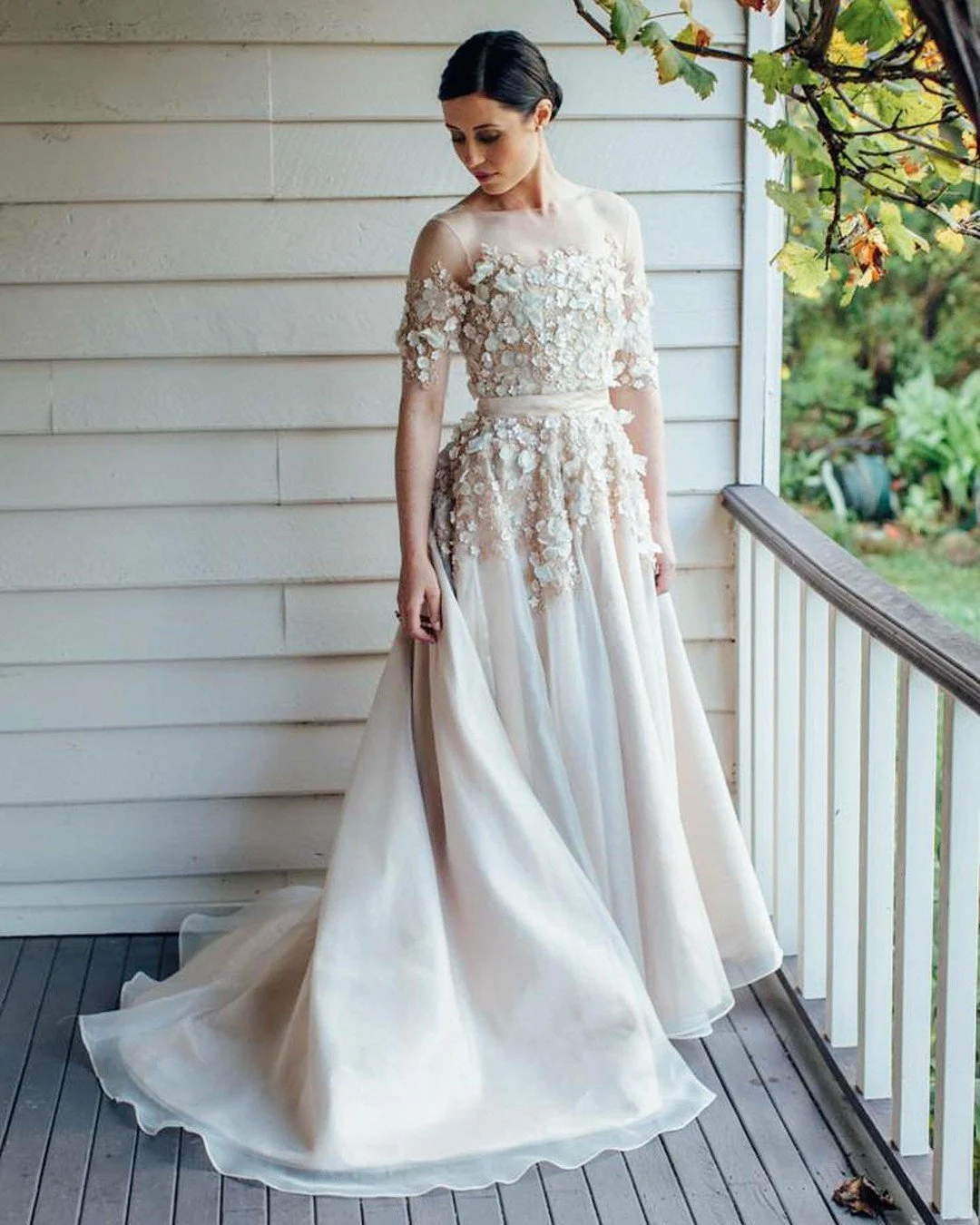 Woman in a wedding dress with floral lace details on a porch with white wooden siding and green foliage outside.