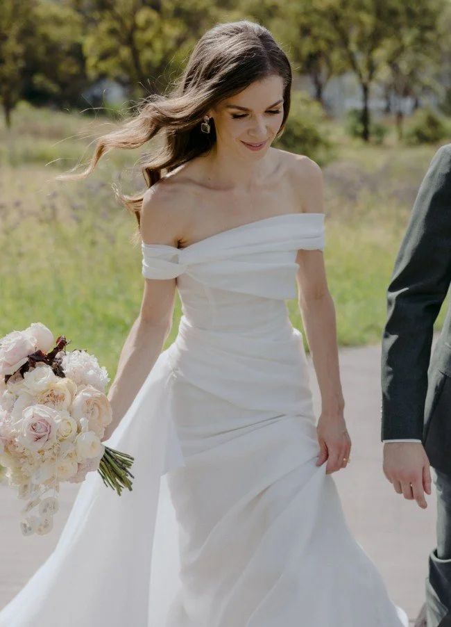 A bride in an off-the-shoulder white wedding gown holding a bouquet of pink and white flowers, walking outdoors near greenery.