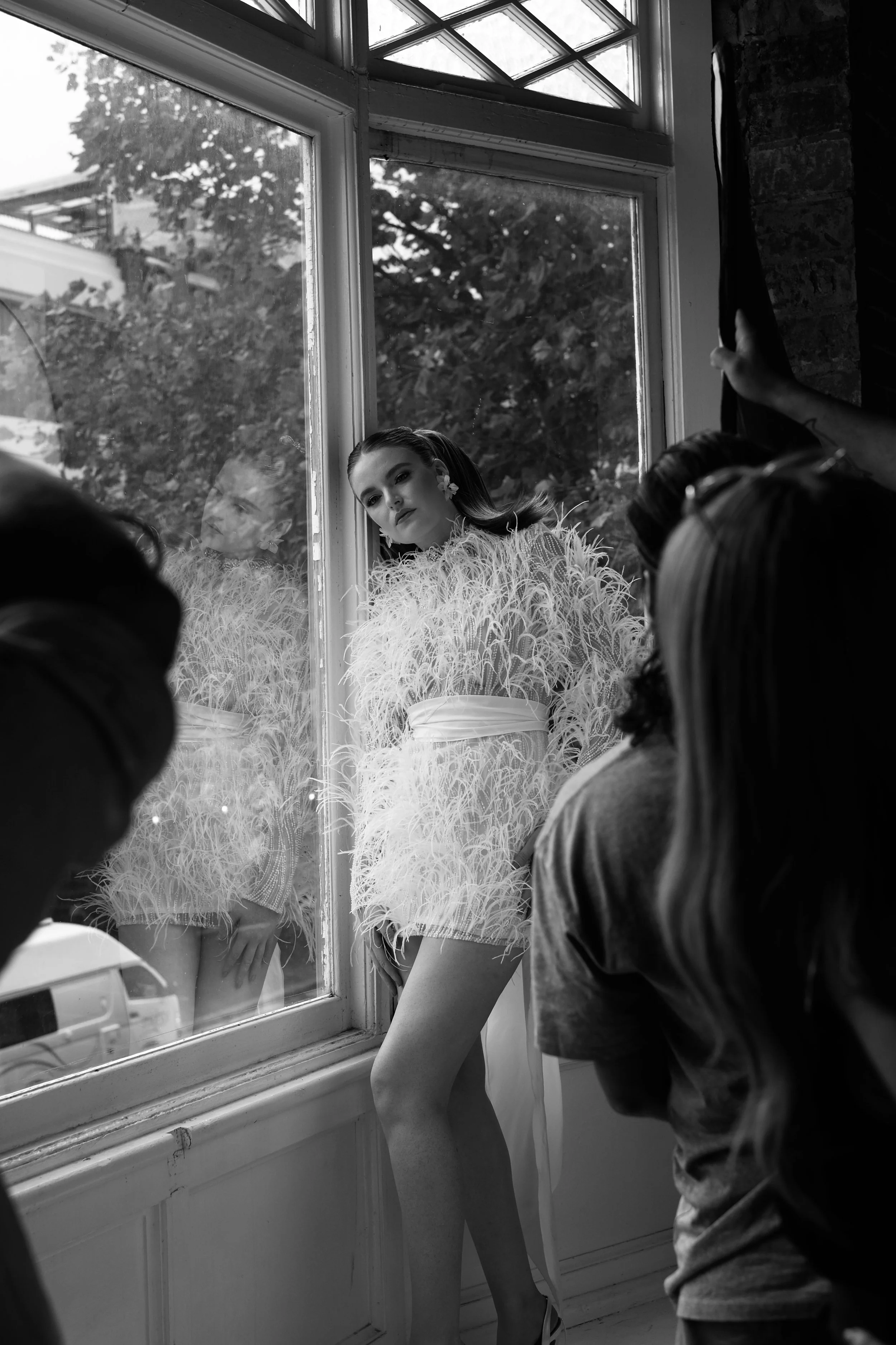 A woman in a feathered dress posing by a large window with trees outside, while a group of people surrounds her, some adjusting curtains.